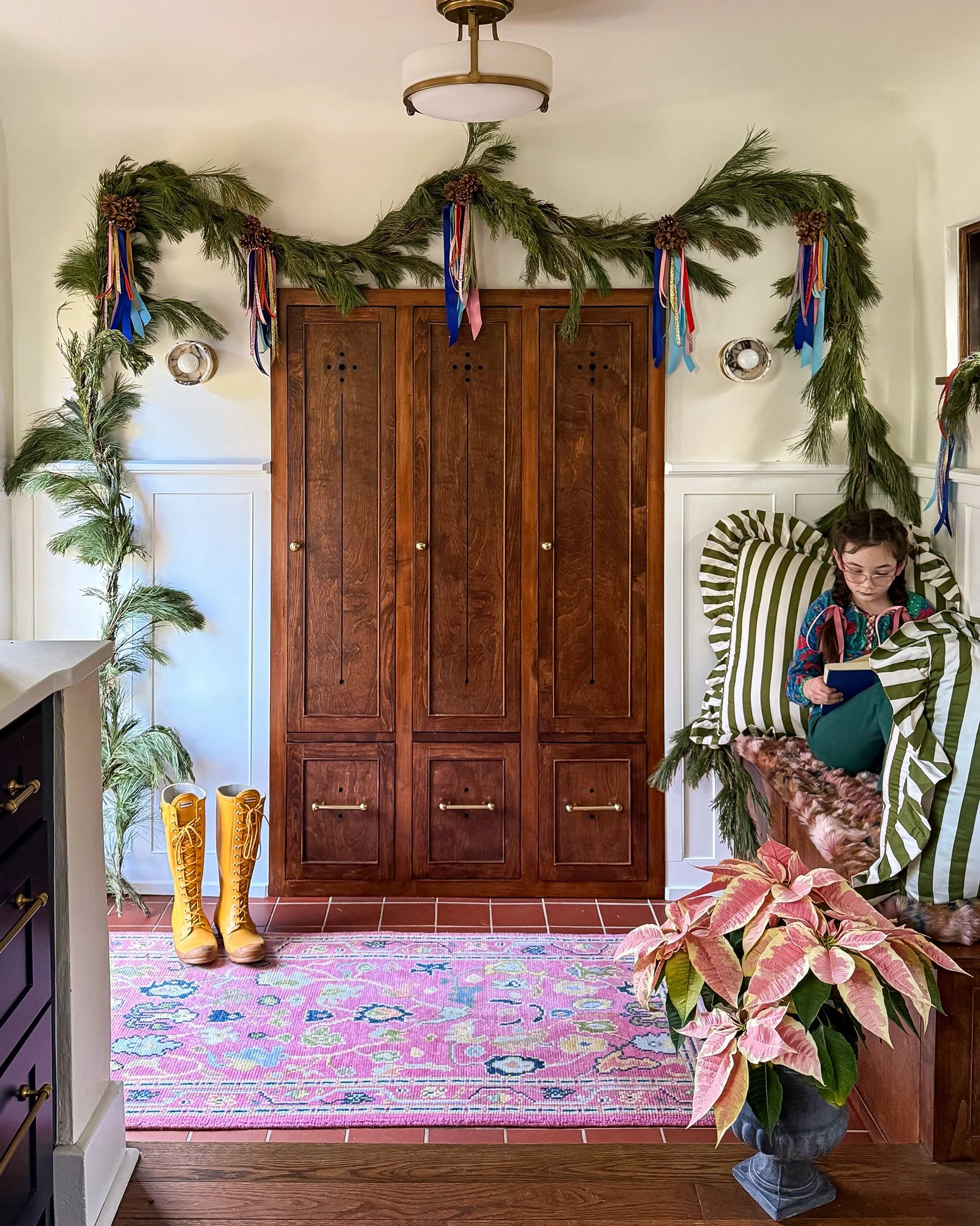 A decorated entryway with a wood door, a pine garland with colorful ribbons hanging above, and a colorful patterned rug on the floor. Yellow snow boots are next to a green leafy plant, and a girl is seated in a green-and-white striped chair reading a