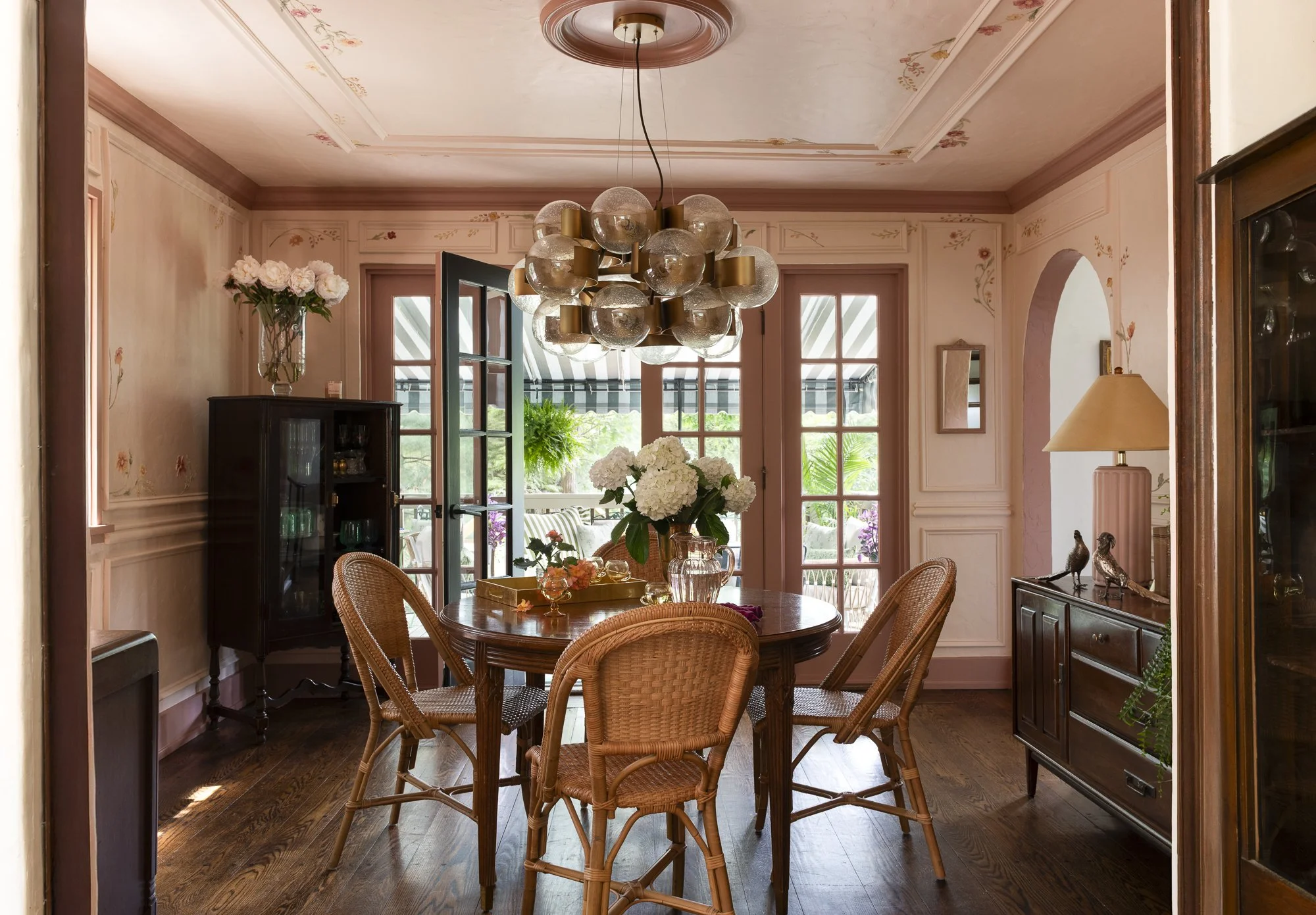 A dining room with a wooden round table and beige wicker chairs, decorated with a vase of white flowers, surrounded by pink-painted walls with floral accents, and a modern chandelier hanging overhead. Large French doors lead to an outdoor patio with 