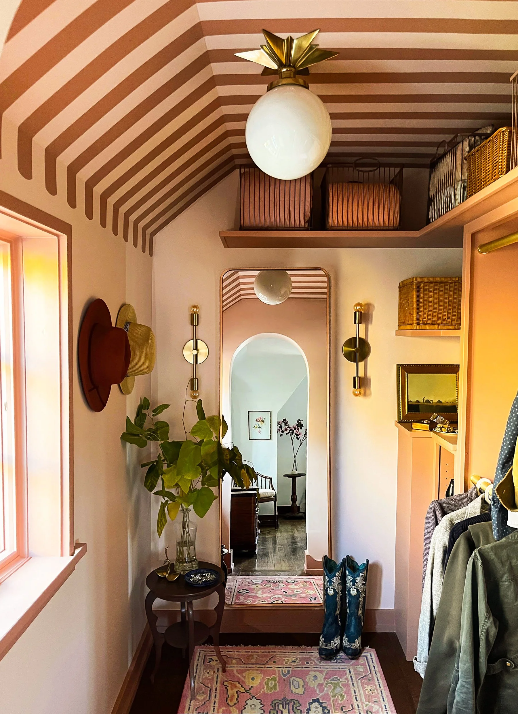 Entryway with pink and brown striped ceiling, wall-mounted lights, a small wooden side table with a glass vase of green plants, a mirror, a rug, and a pair of cowboy boots. There are storage baskets on a shelf above, and clothes hanging on the right 