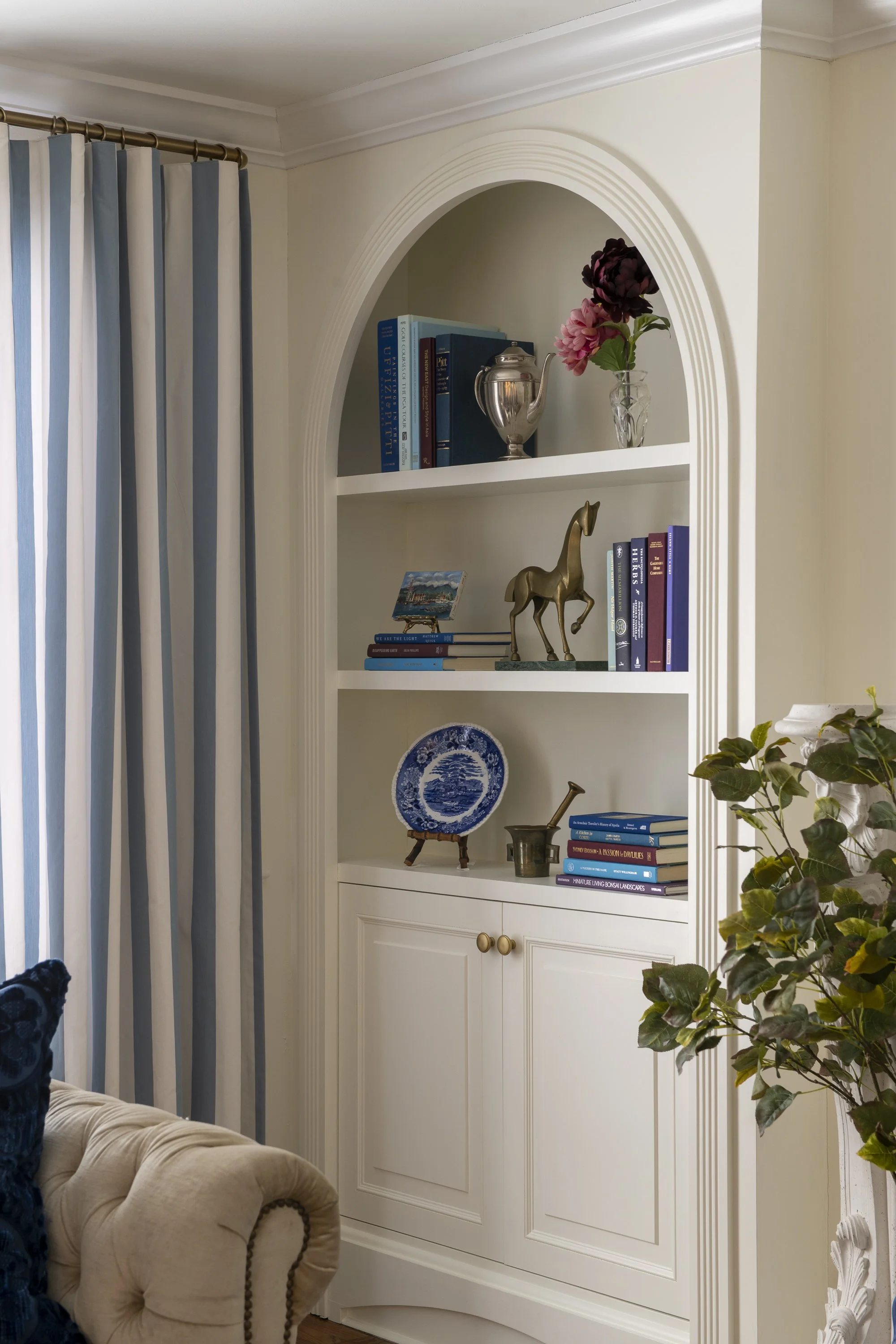 Interior of a living room featuring a built-in white bookcase with decorative molding, holding books, a silver teapot, and a brass giraffe figurine. To the left, there are blue and white striped curtains, and to the right, a green leafy plant.