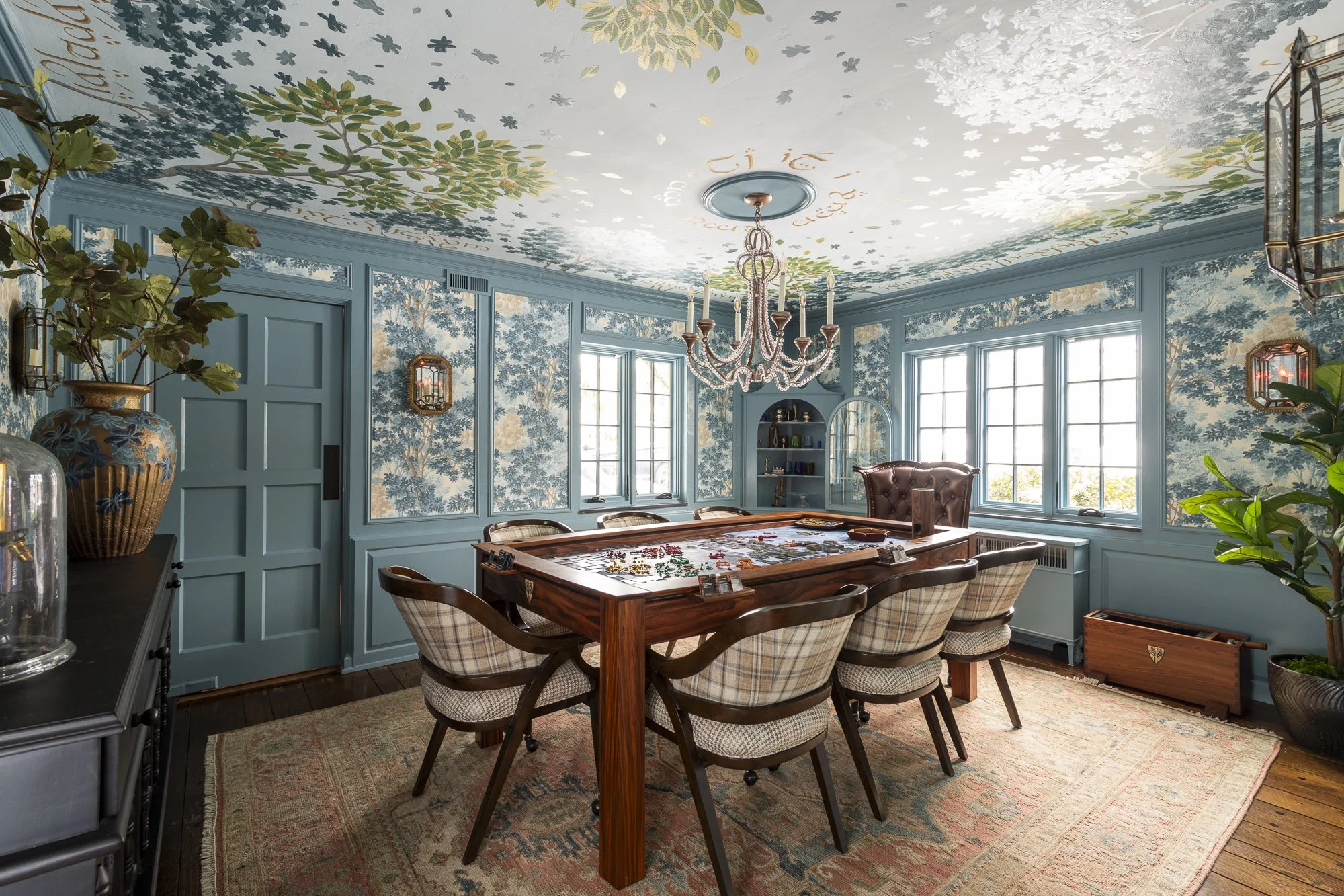 A room with light blue walls and intricate murals featuring trees and foliage, large windows allowing natural light, a large painted wooden table with chairs around it, and a chandelier hanging from the ceiling. There are decorative vases and plants,
