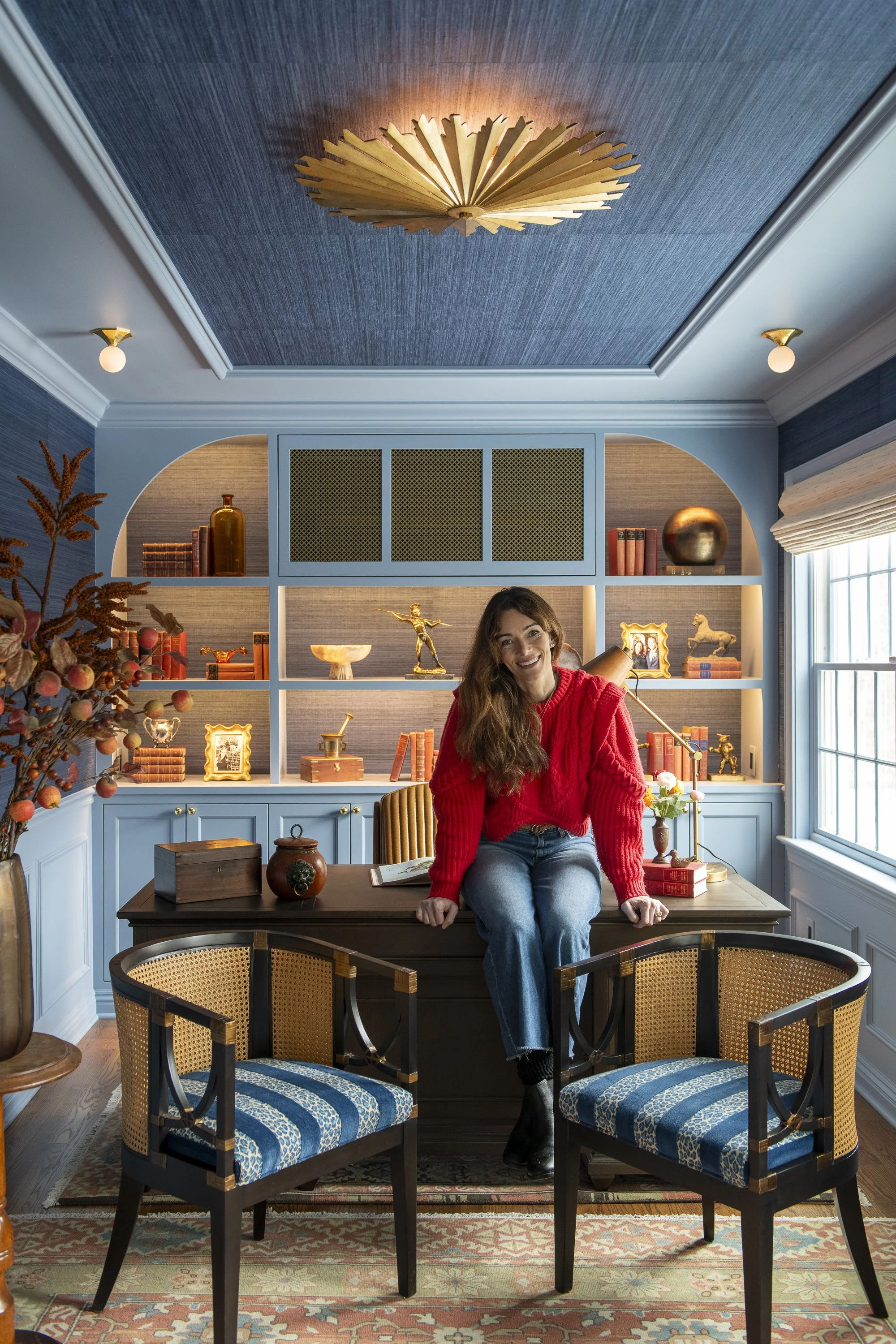 A woman with long brown hair, wearing a red sweater and blue jeans, sitting on a dark wooden table in a home office or library. She is smiling, with one hand resting on the table and the other on her lap. The room has a large built-in bookshelf with 