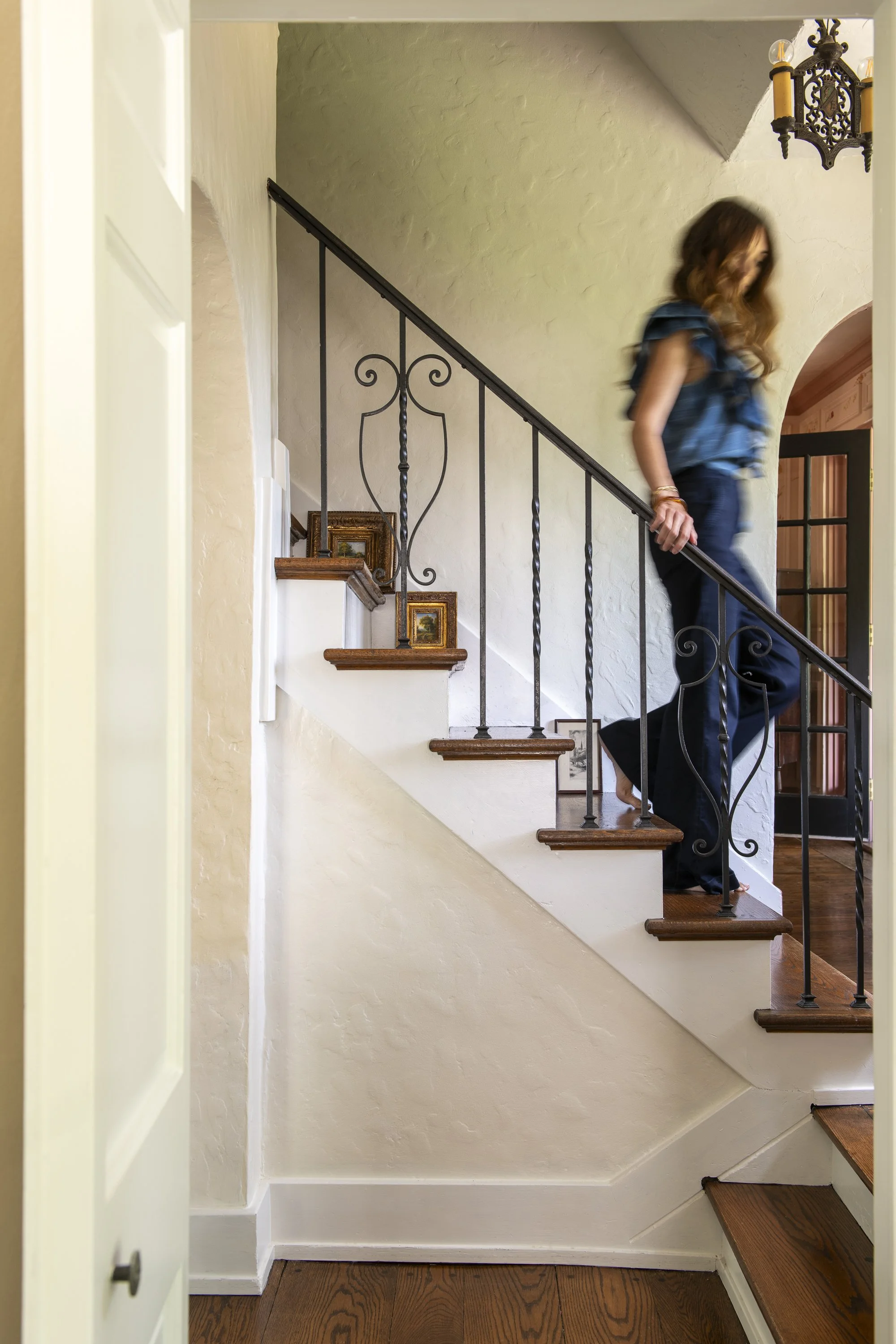 Inside view of a staircase with dark wooden steps, black wrought iron railing, white textured walls, small framed pictures on a ledge, and a woman with wavy hair wearing a blue top and dark pants descending the stairs.