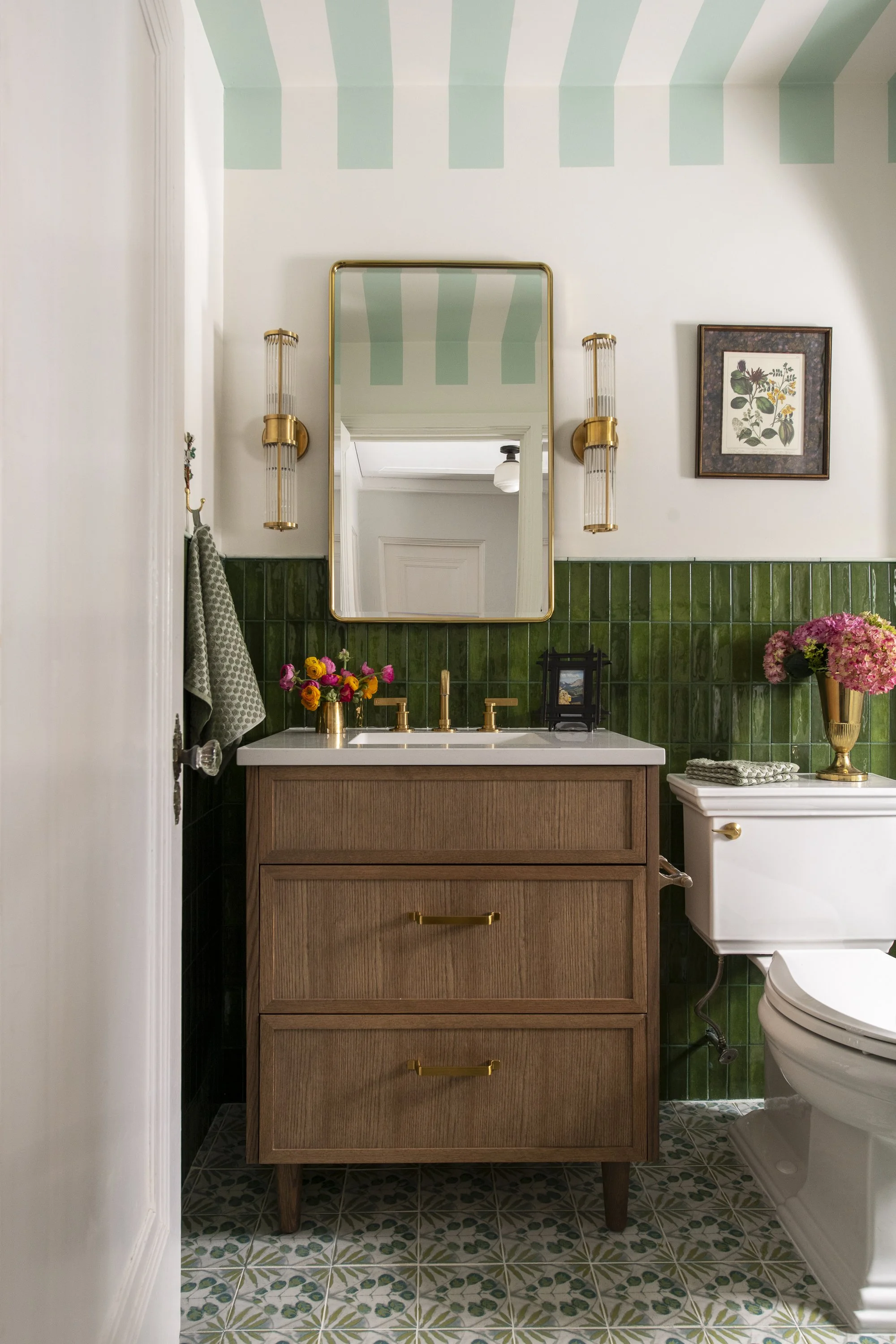 Bathroom with a wooden vanity and white countertop, green tiled backsplash, framed botanical print on the wall, gold and glass wall sconces, a mirror with a gold frame, flowers in vases, and a toilet to the right with a white side table beside it.
