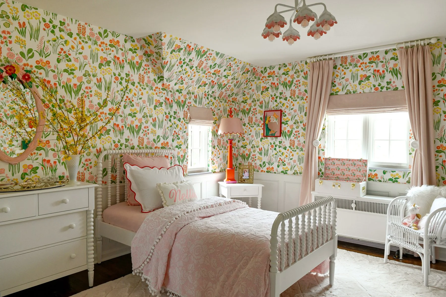 A bedroom with floral wallpaper, a white bed with pink and white pillows, a white dresser with a vase of yellow flowers, a white nightstand with a pink lamp, a window with curtains, and a white wicker chair with stuffed animals.