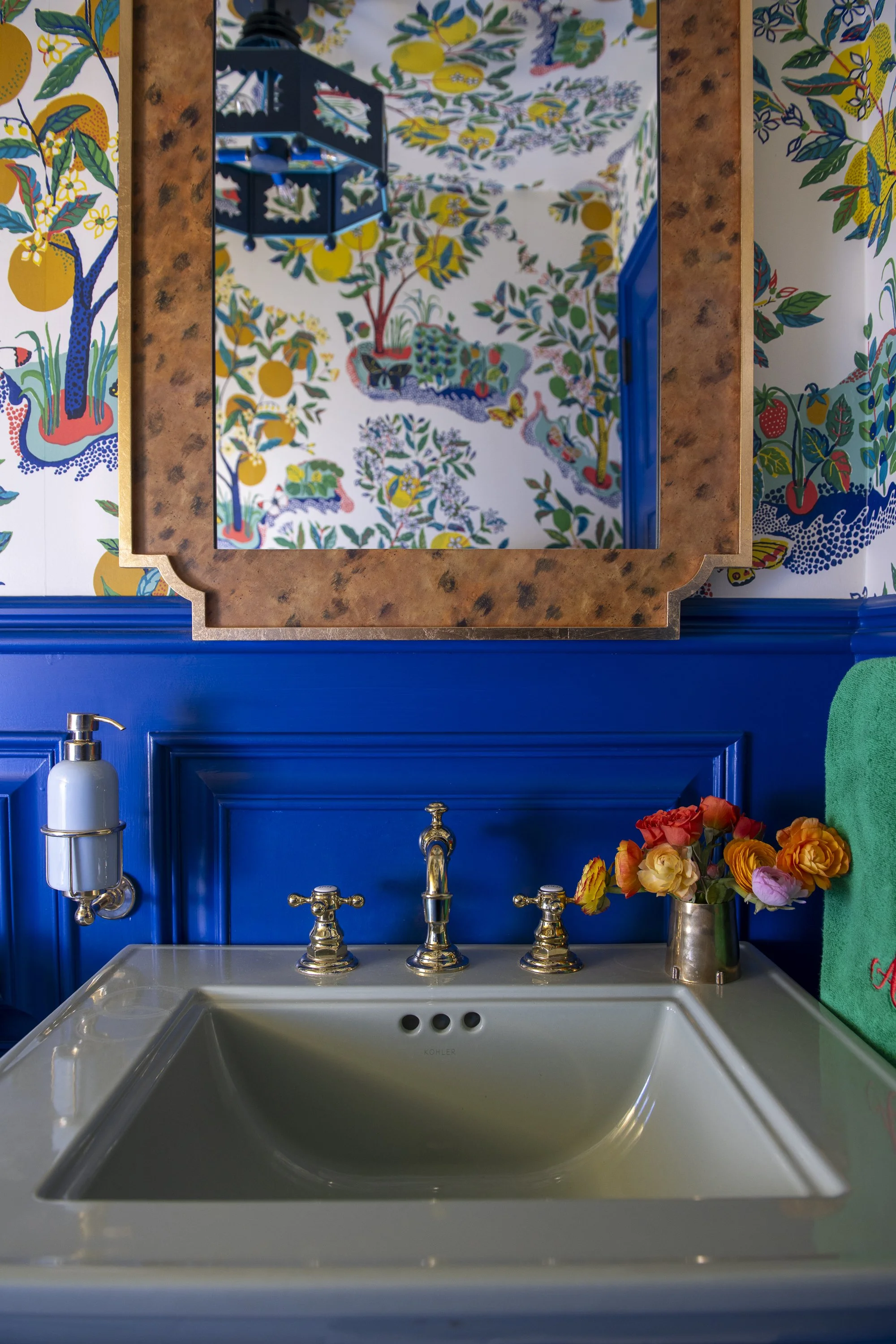 A bathroom sink with a blue cabinet, floral patterned wallpaper, a mirror, a faucet, and a flower arrangement in a metallic vase.