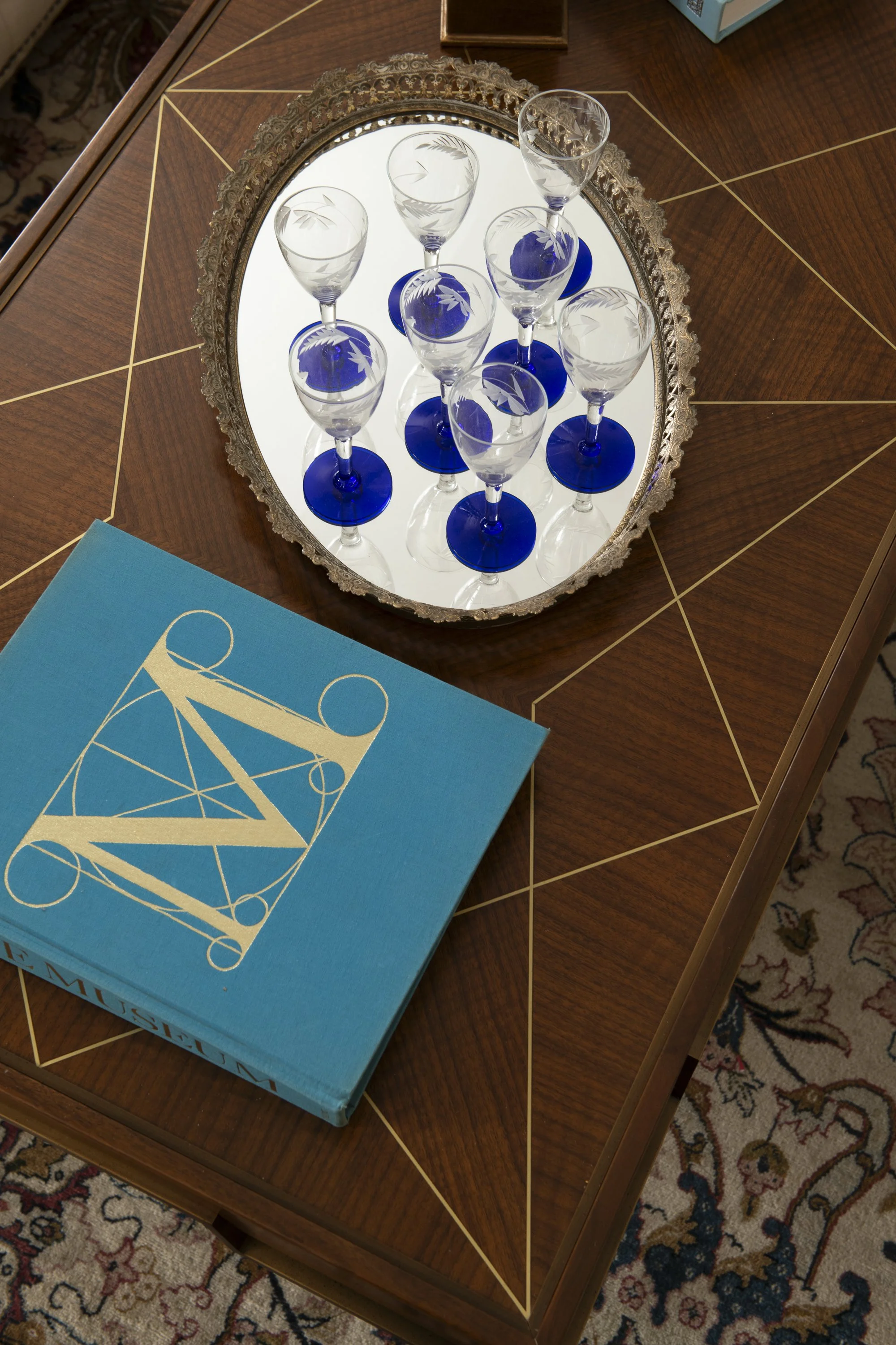 A wooden table with gold geometric inlay, reflecting an oval mirror on top, holding ten wine glasses with blue bases and etched patterns, and a blue box with a geometric design and the text 'L Müller' on the bottom edge.