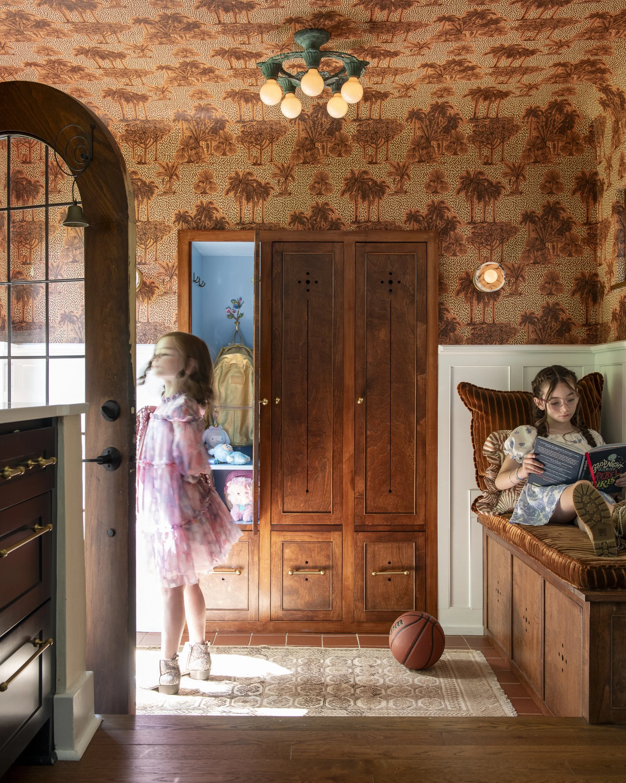 A young girl is standing in a kitchen near a doorway, with sunlight coming through. She is wearing a pink and white dress with sparkly shoes, and has long hair. On the floor in front of her, there's a basketball. To the right, a girl with glasses is sitting on a cushioned chair, reading a book, with her hair in braids and wearing a patterned dress. The kitchen has wooden cabinets, a tiled floor, and a patterned rug. There is a vibrant wallpaper with a tropical tree and animal design, adding a vintage touch to the room.