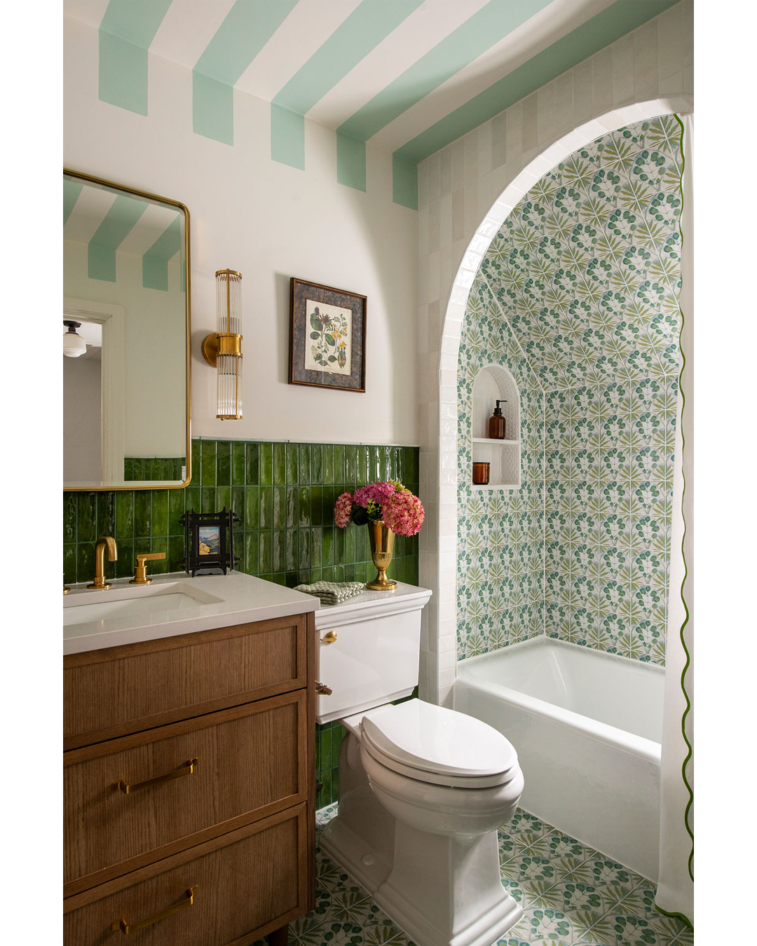 A bathroom with a green and white color scheme, featuring a wooden vanity with gold handles, a white toilet, a white bathtub with a patterned tiled wall, a large mirror with a gold frame, framed botanical art, a gold wall sconce, and a vase with pink