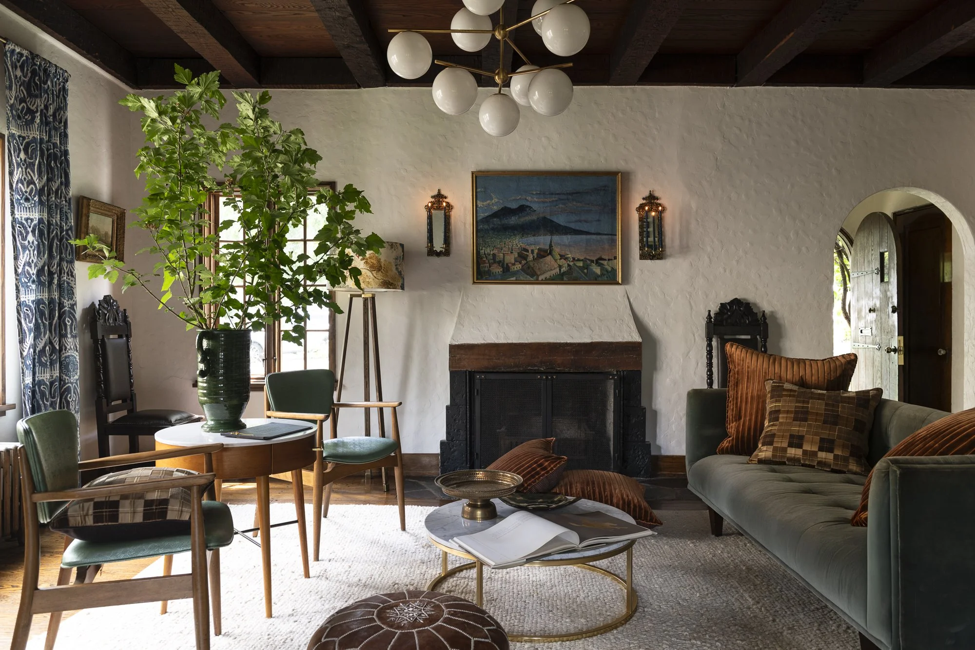 Living room with a white textured wall, dark wooden beam ceiling, and a modern chandelier with white globes. There is a green plant in a dark vase, a fireplace, framed artwork, and a mix of chairs and a sofa with plaid and solid pillows. A window and