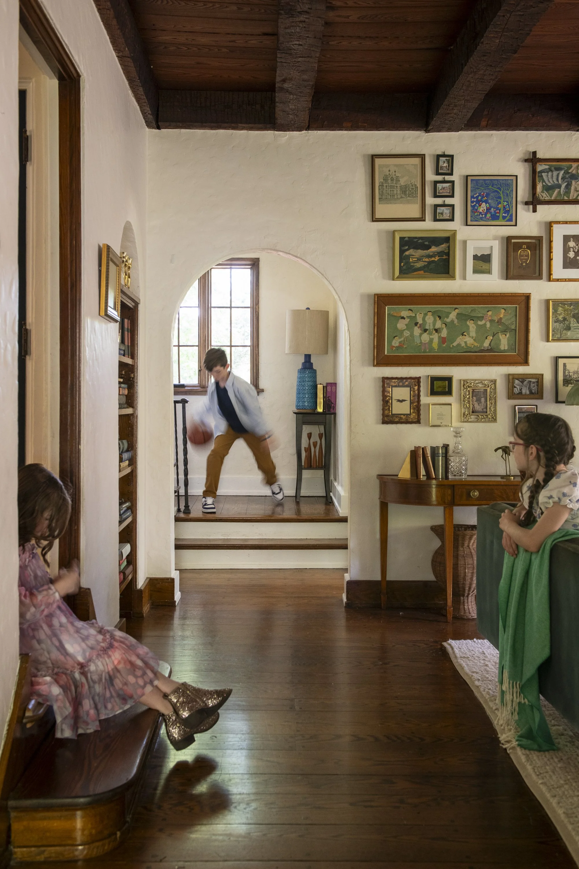 A young boy in a blue shirt and tan pants is playing basketball in a living room with white walls, wooden floors, and a high wooden ceiling with exposed beams. Several children and a woman are watching him. The room is decorated with framed artwork, 