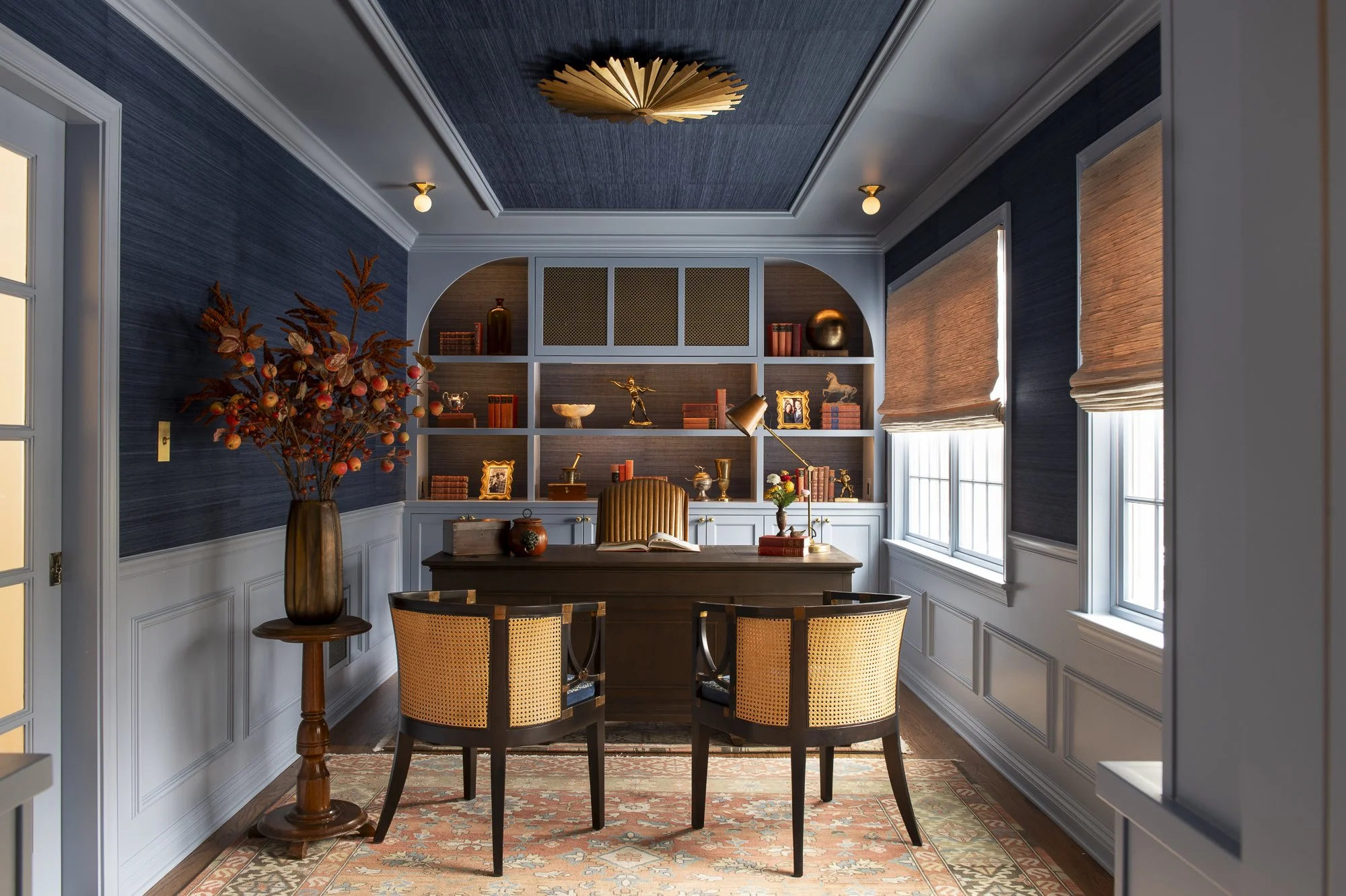 A home office with blue walls, white trim, a wooden desk with a chair, a large bookshelf above the desk with decorative objects, books, and pictures, a window with Roman shades, and a floor rug. A tall vase with branches and berries is on the left si