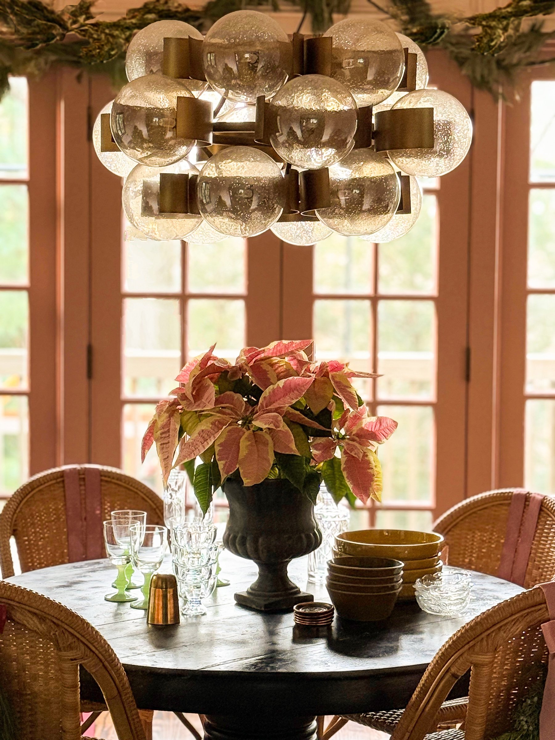 A round dining table decorated with a large potted poinsettia centerpiece, surrounded by wicker chairs, with a chandelier hanging above and a window with wooden framing in the background.