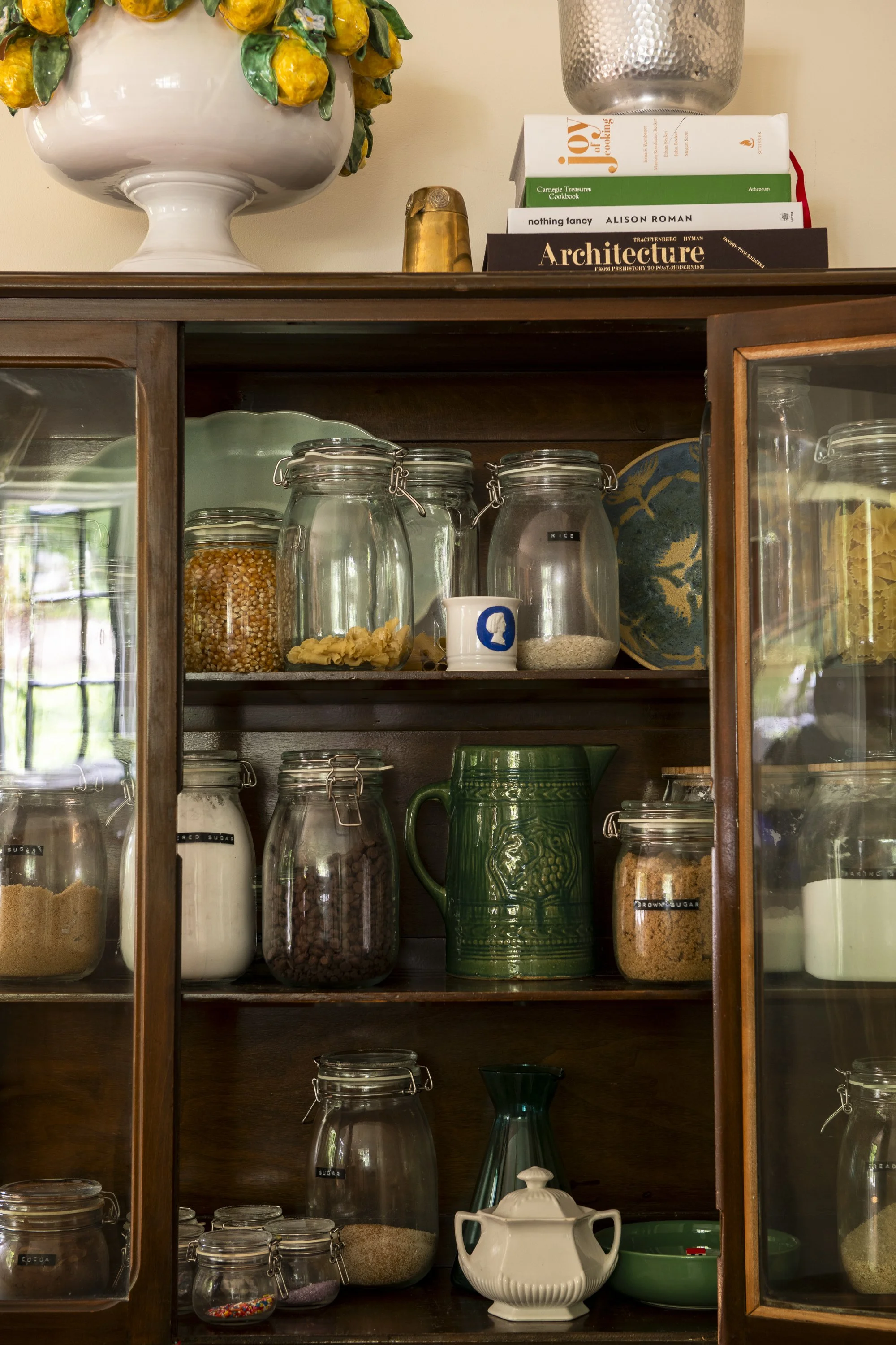A wooden kitchen cabinet filled with glass jars of baking ingredients such as sugar, flour, chocolate chips, and rice. On top, there is a large decorative ceramic bowl with lemons, stacked cookbooks, and a metal vase.