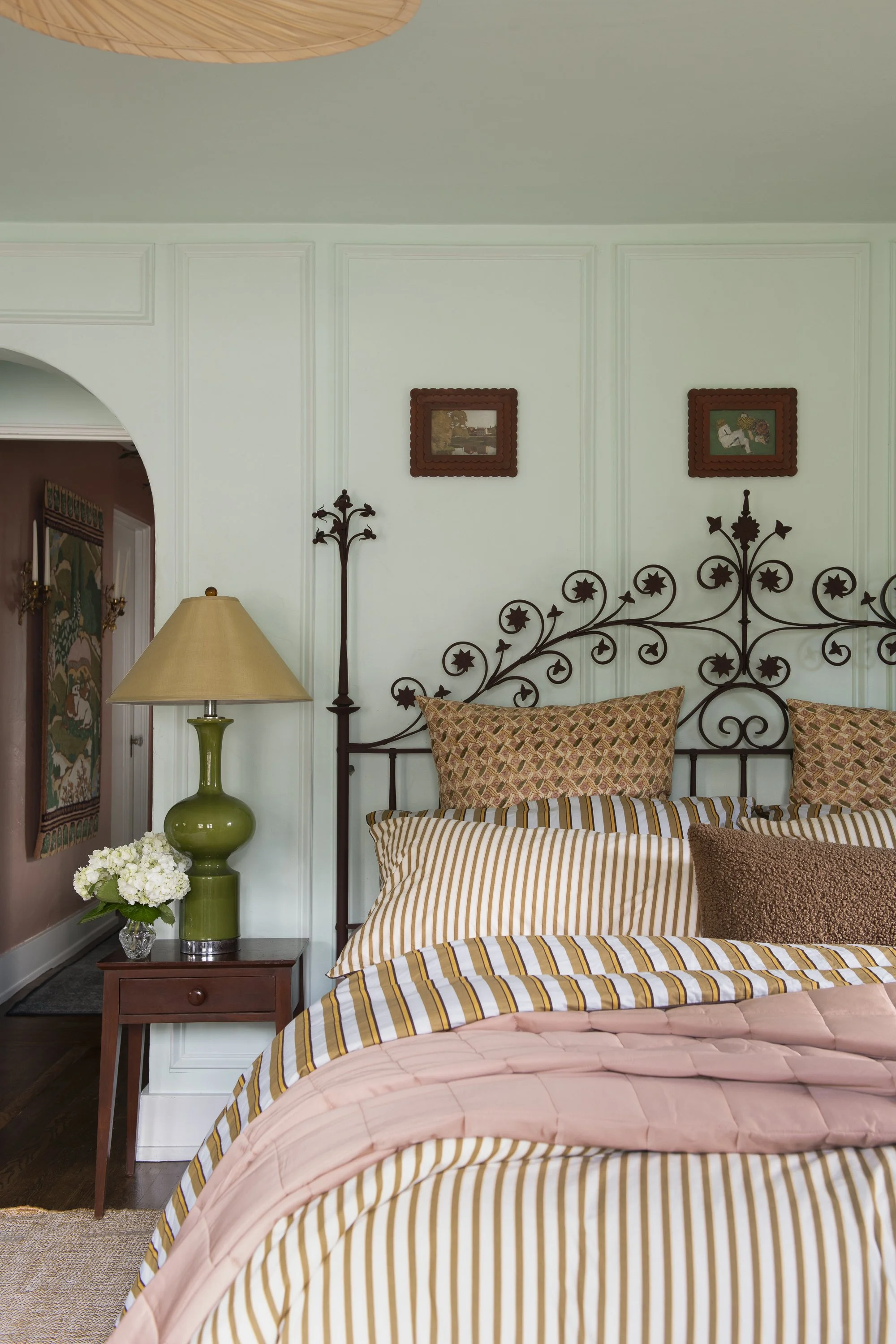 A bedroom with a black metal bed frame featuring decorative star and swirl patterns, a striped duvet cover in shades of gold, white, and pink, and matching pillows. There is a wooden side table with a green ceramic lamp and a glass vase of white flow