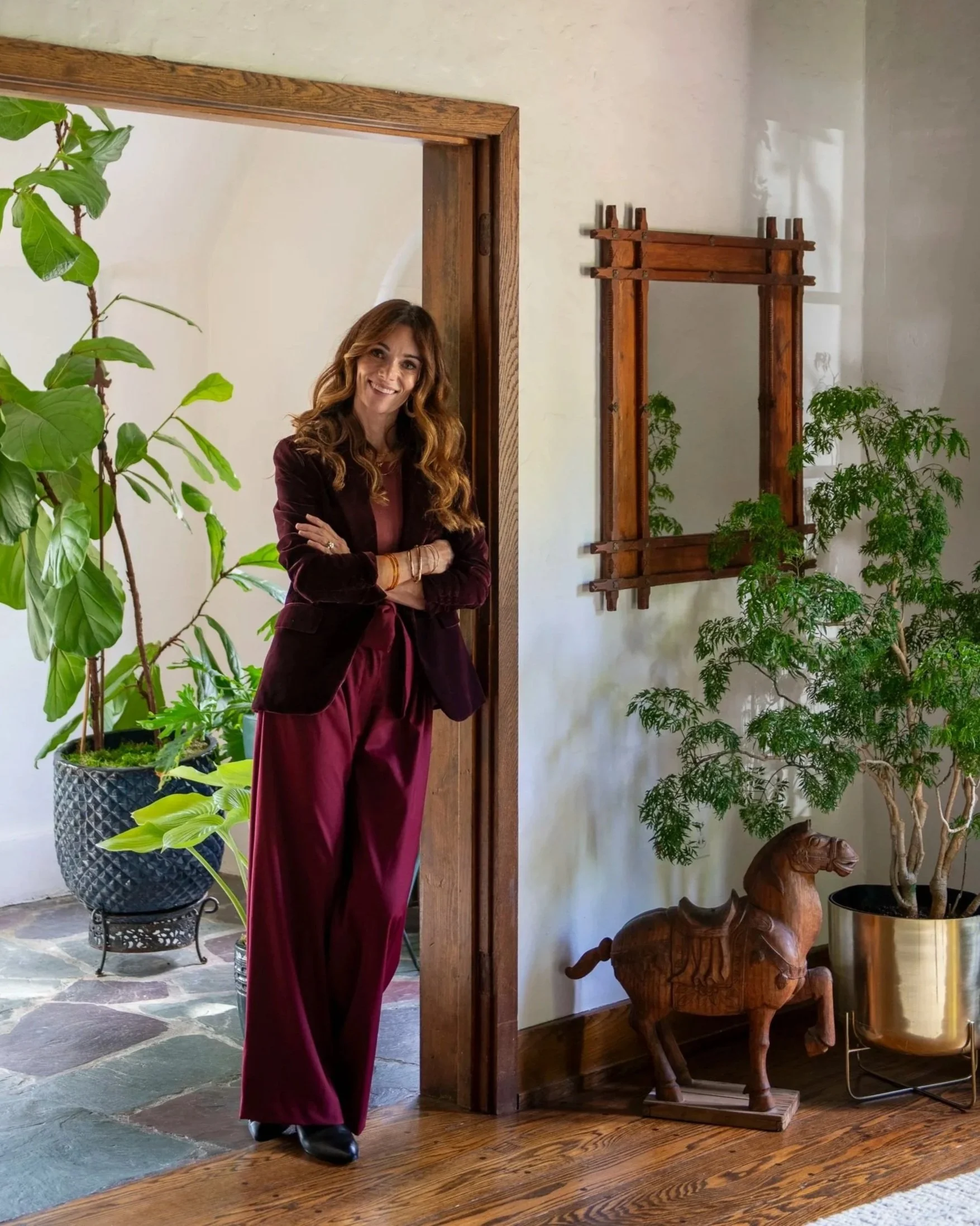 A woman with long wavy brown hair standing indoors. She is smiling, wearing a maroon velvet blazer and matching flowy pants. She is crossing her arms and wearing multiple bracelets. The background features large green potted plants, a wooden door frame, a carved wooden elephant statue, and a wooden sculpture of a lion on a stand.