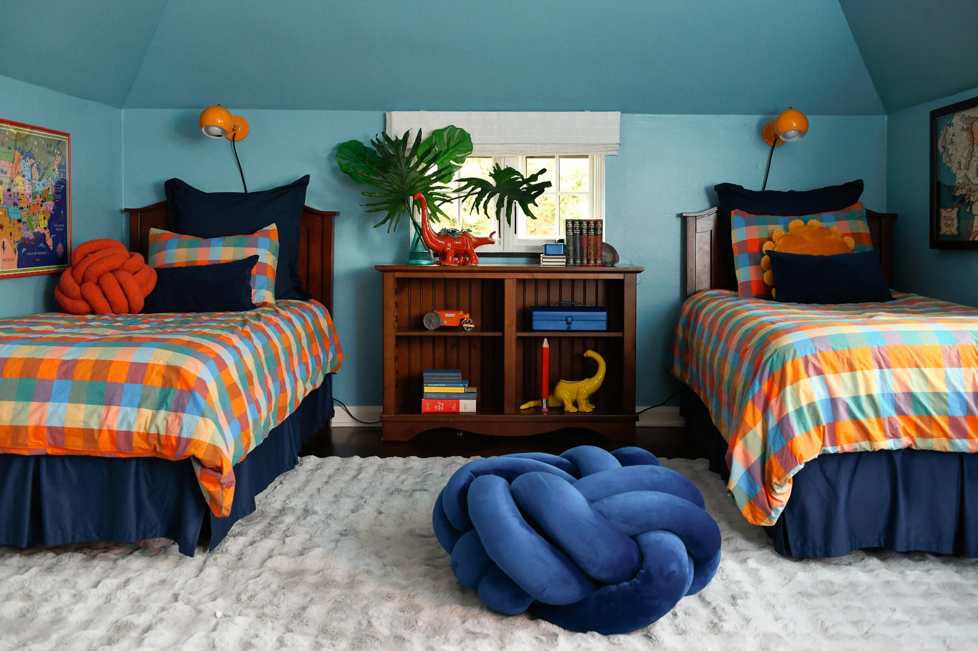 A bedroom with two twin beds with colorful checkered bedding, navy blue pillows, and decorative pillows. A wooden nightstand between the beds holds a red dinosaur-shaped lamp, large green leaves in a vase, and books. The room has two orange wall-moun