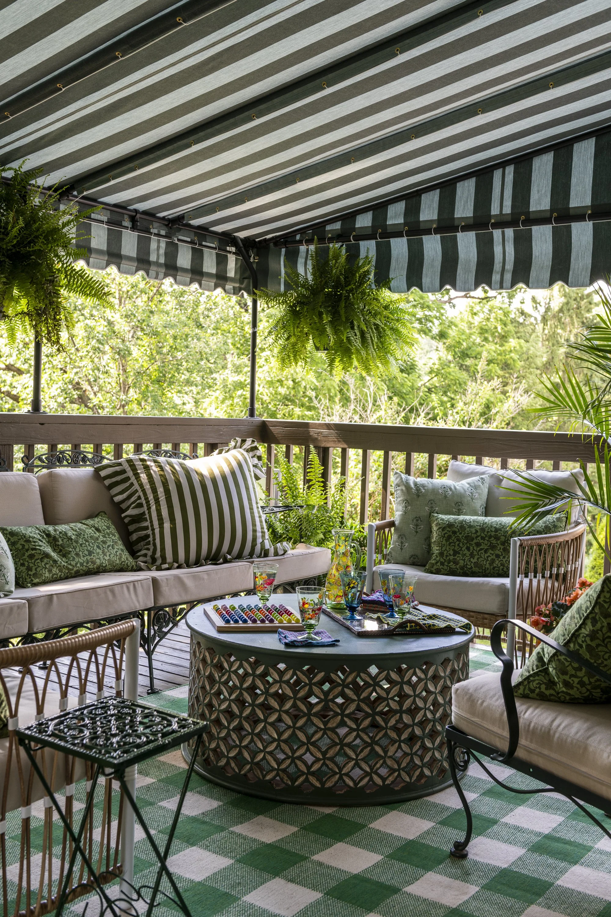 A cozy outdoor patio with a green and white checkered rug, overlooking lush greenery, furnished with a cream-colored sofa and armchair adorned with green patterned pillows, a round metal table with glassware, colorful beads, and a tray, surrounded by