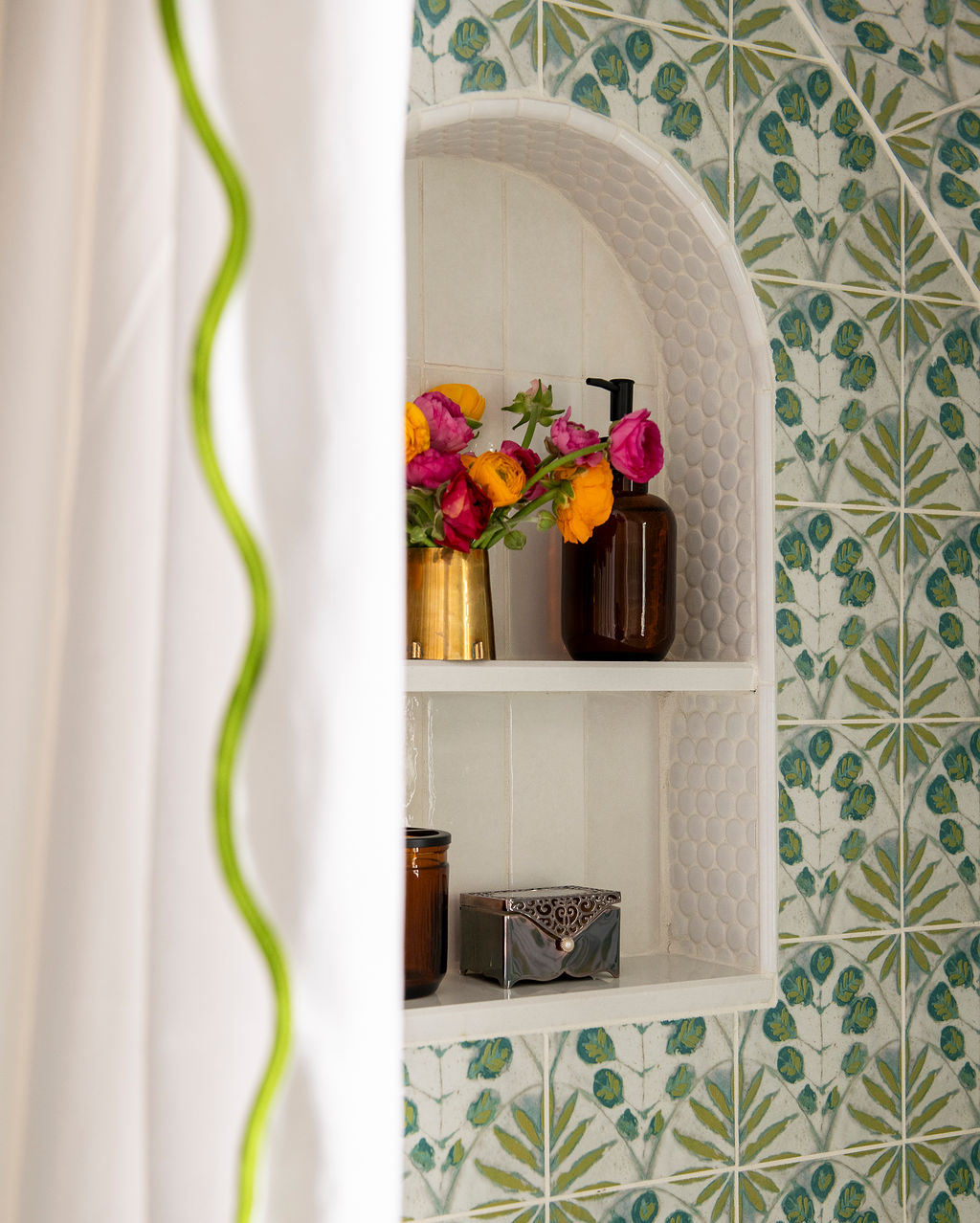A bathroom shelf with a bouquet of pink and yellow flowers in a gold vase, a brown soap dispenser, and small containers, set against a wall with patterned tiles.
