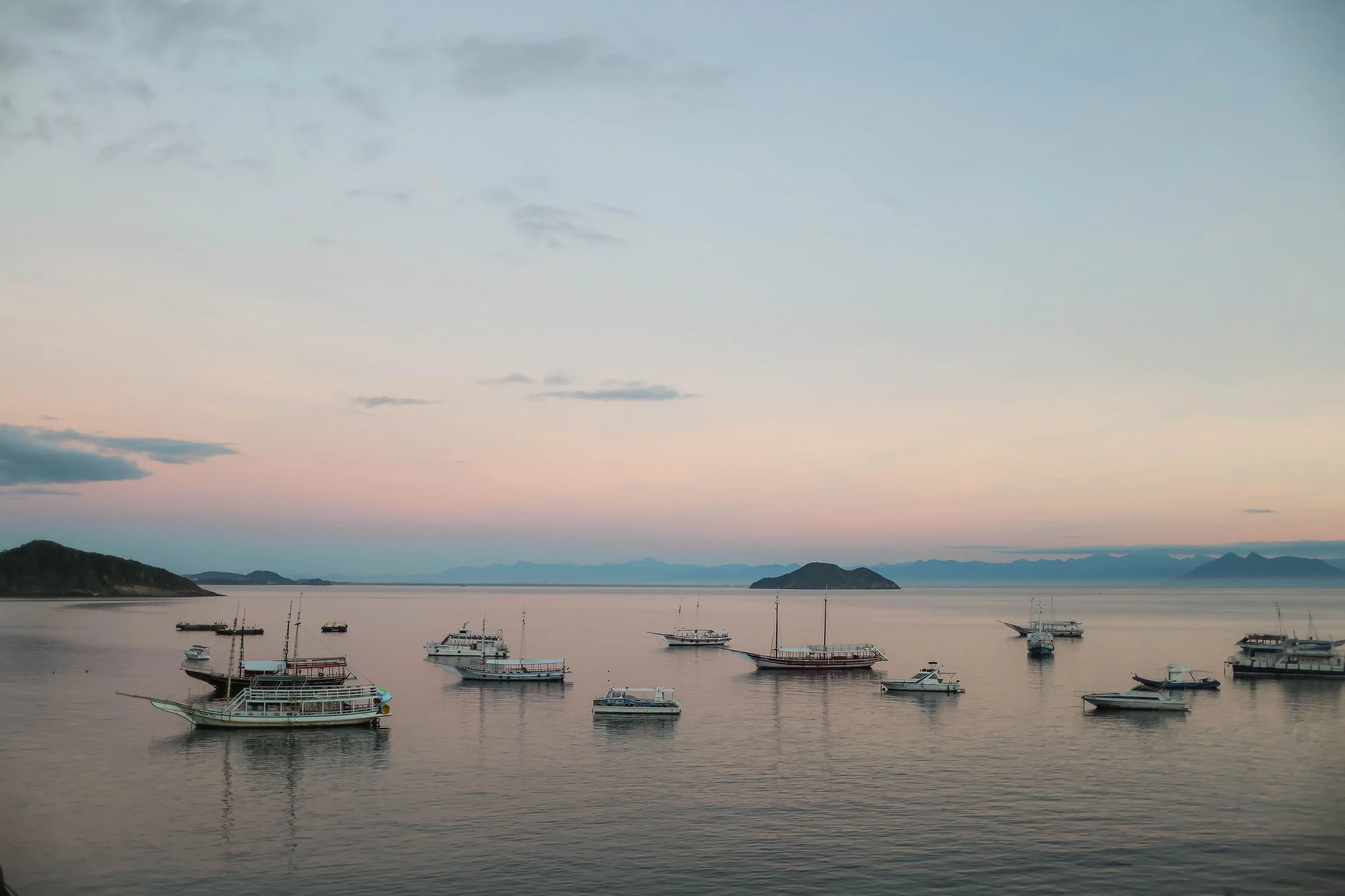 A serene harbor scene at dusk with several boats docked on calm water, overlooking a mountainous landscape under a pastel-colored sky with scattered clouds.