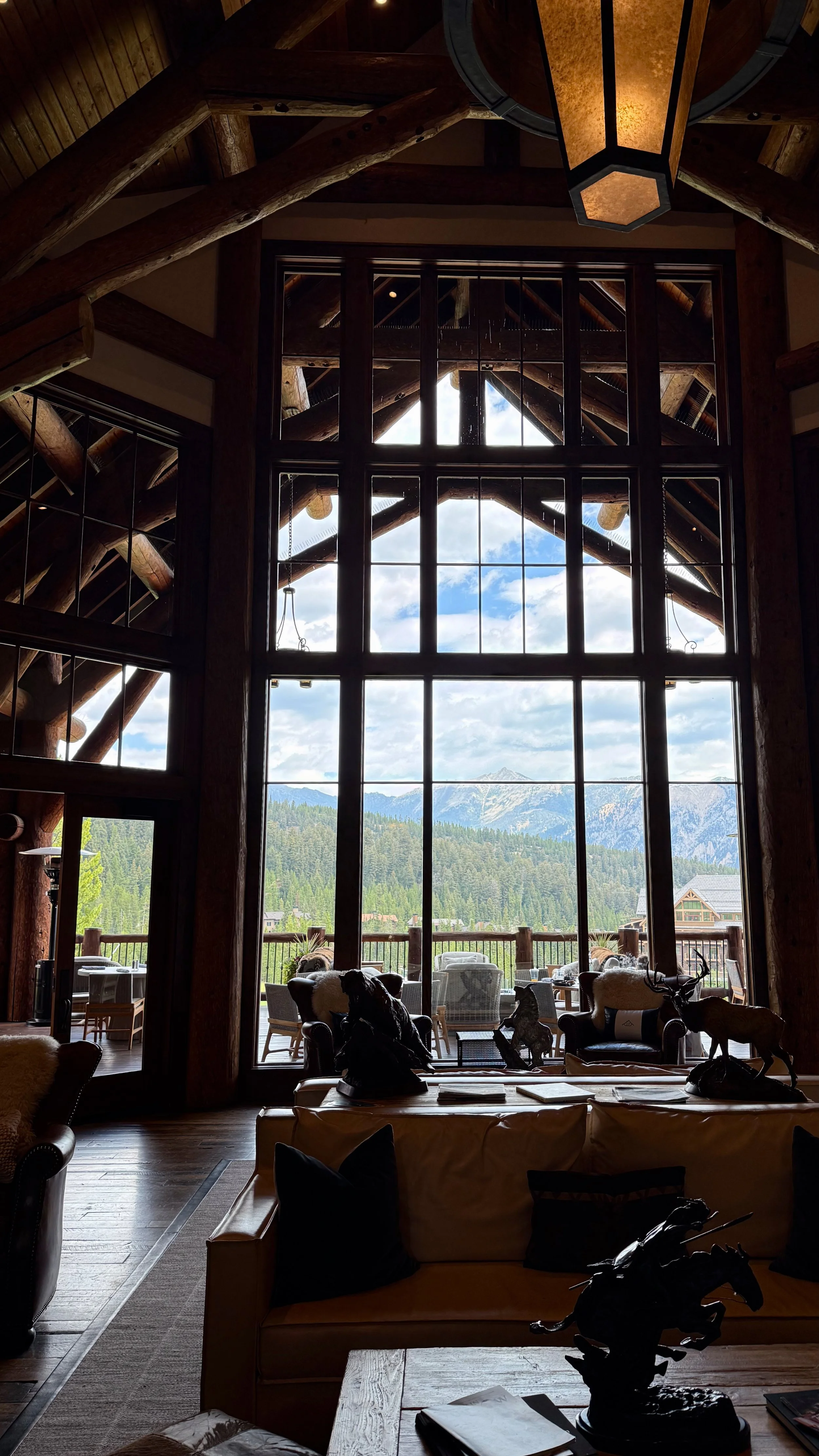 Inside a rustic-style lodge with large windows showing a mountain landscape. The interior features wooden beams, a hanging lantern light, and various sculptures on tables and furniture.