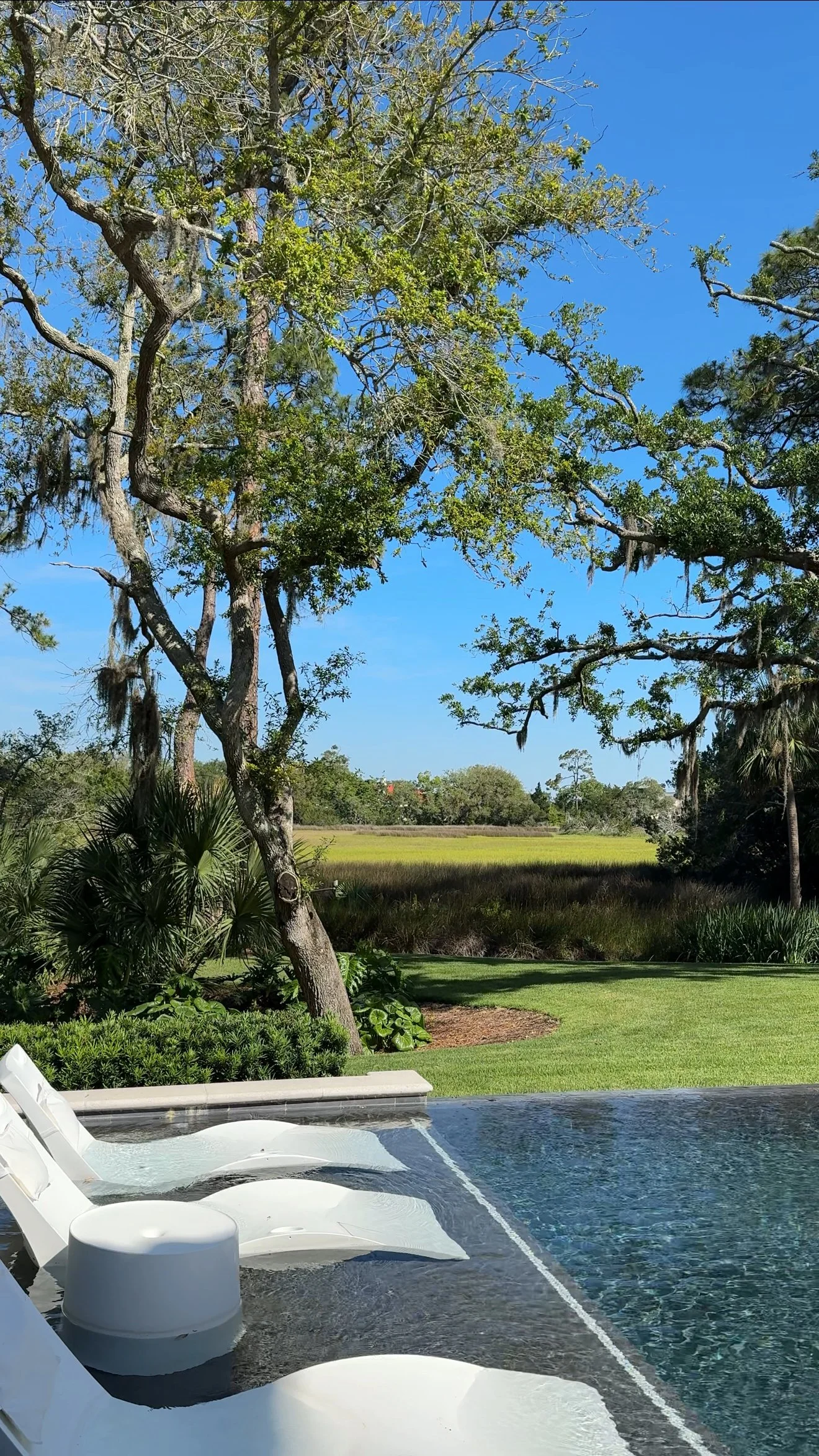 View of a backyard with a swimming pool, lounge chairs, lush green grass, trees, and a clear blue sky in the background at Sea Island Resort in Georgia.