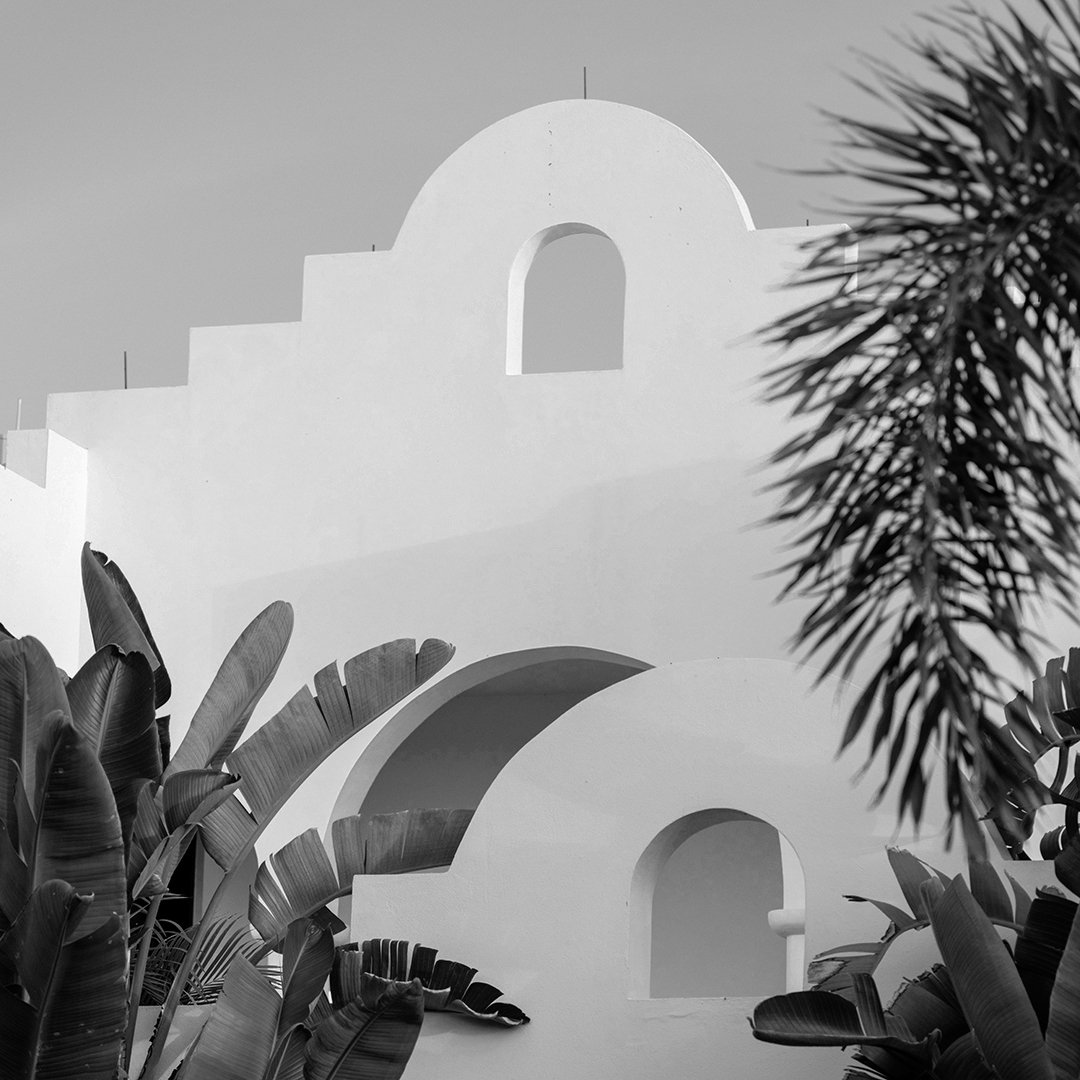 Black and white photo of a white building with arched windows and a stepped, curved roofline, partially obscured by tropical plants.