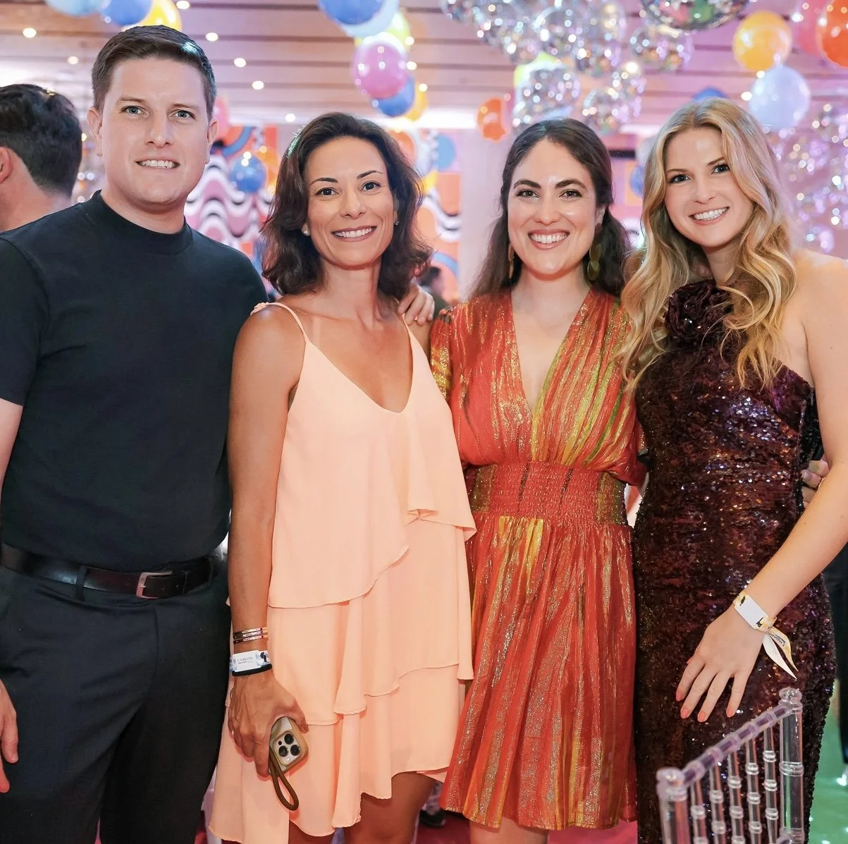 Four people smiling at a festive event with colorful balloons and decorations in the background.