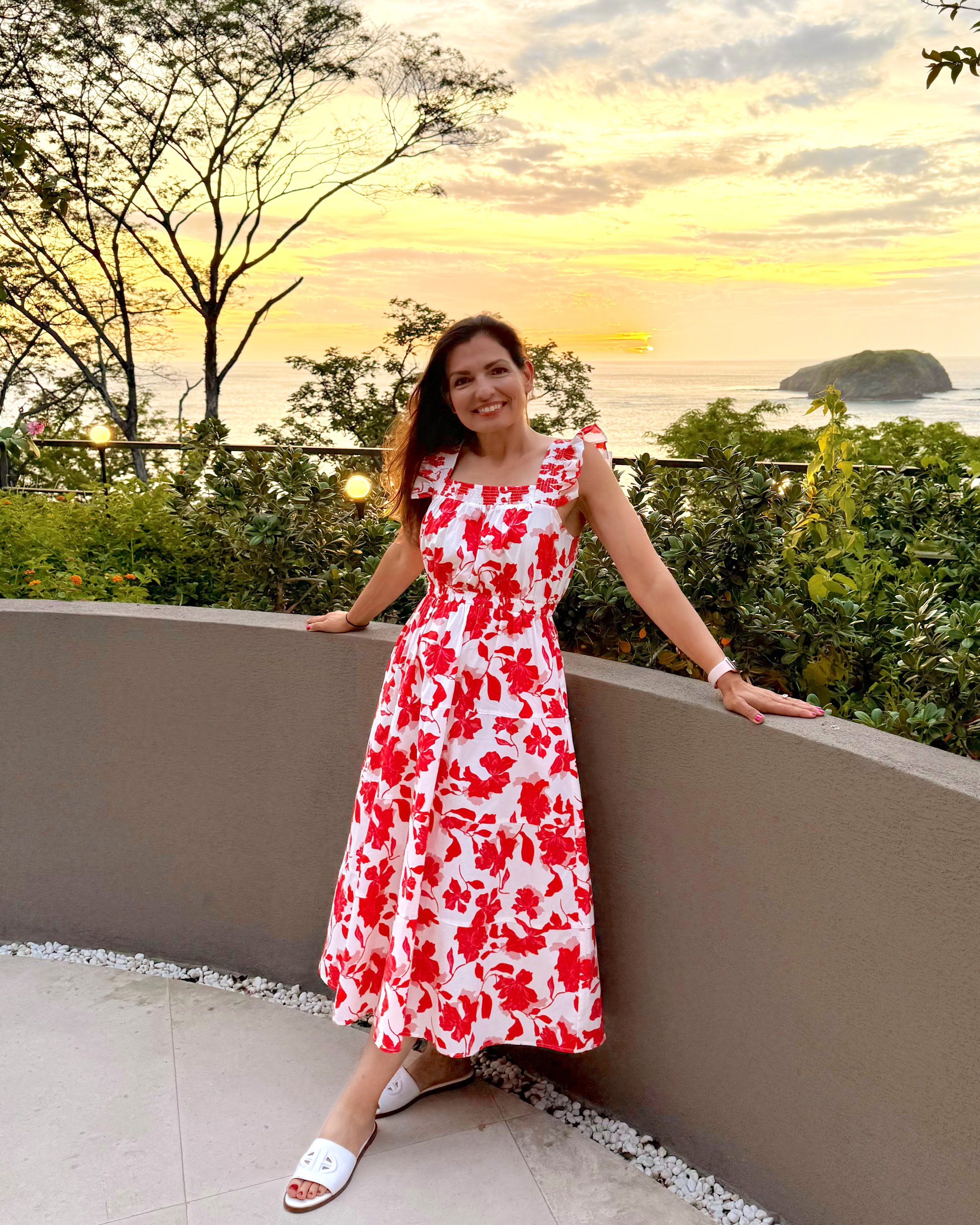 A woman in a red and white floral dress smiling and leaning against a railing on a balcony overlooking the ocean at sunset, with trees and a small island in the background.