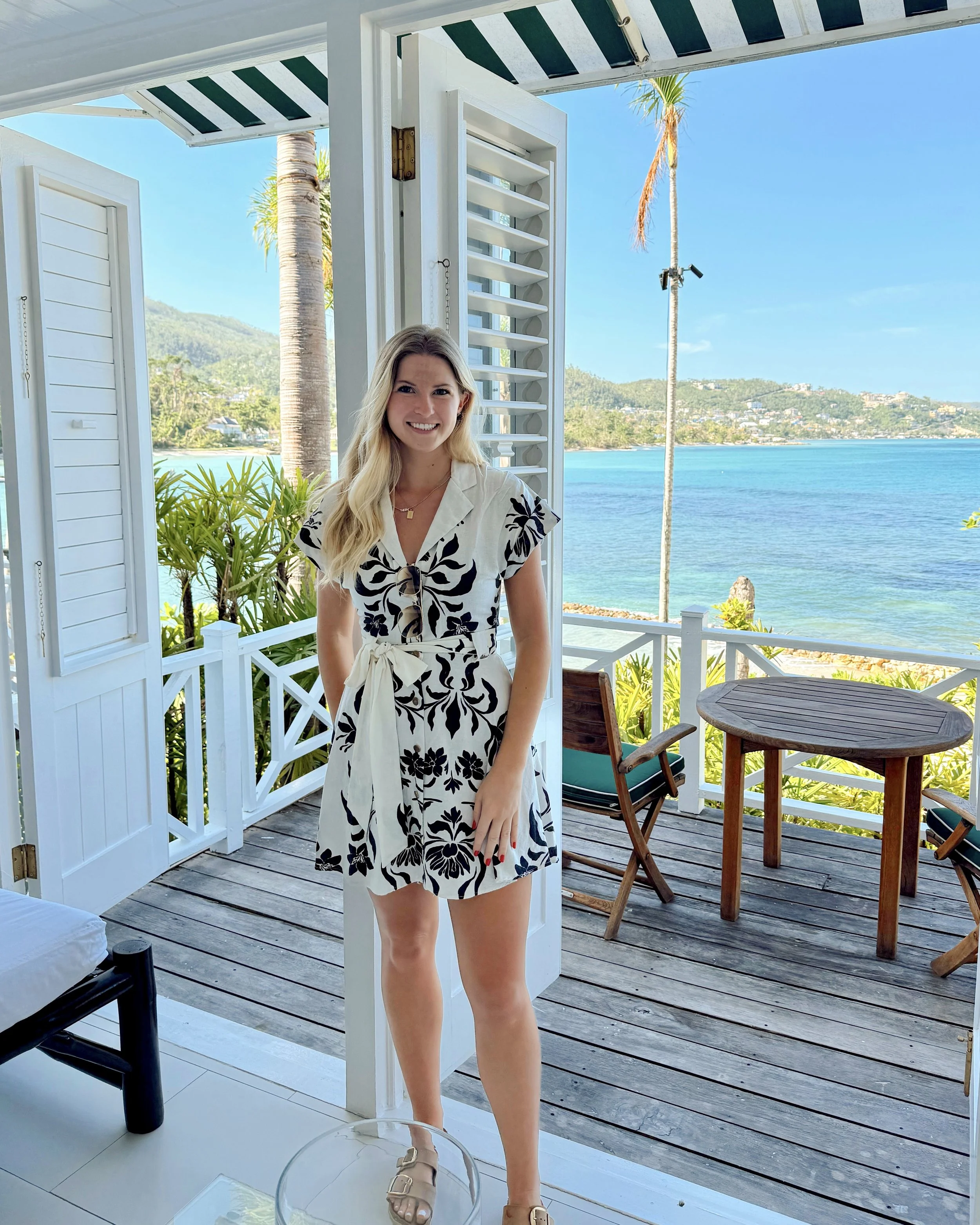 A woman with long blonde hair, wearing a white dress with black floral patterns, standing on a balcony overlooking a body of water and lush, green hills in the distance. The balcony has white wooden shutters and outdoor furniture, with some plants visible.