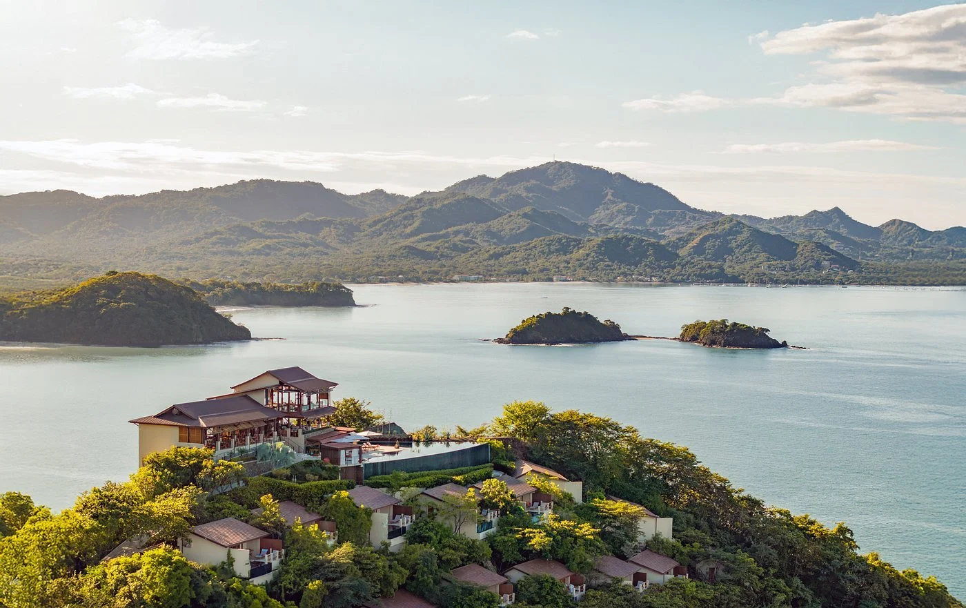 A scenic view of a lake with small islands and misty mountains in the background, with a residential area with houses and lush greenery in the foreground.