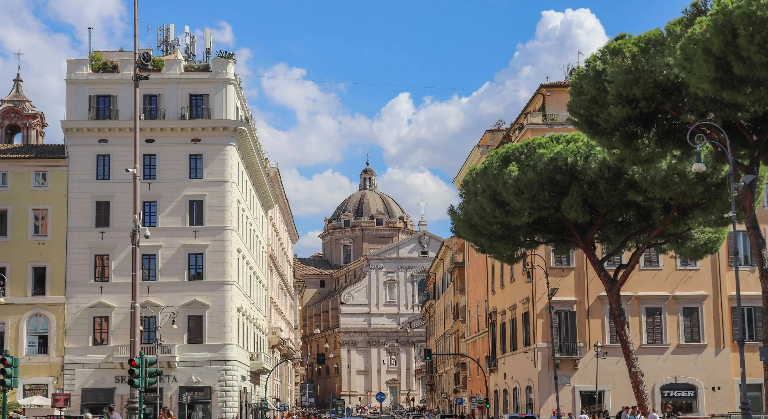 City street scene with multi-story buildings, a domed church in the background, large trees, traffic lights, and people on the street under a partly cloudy sky.