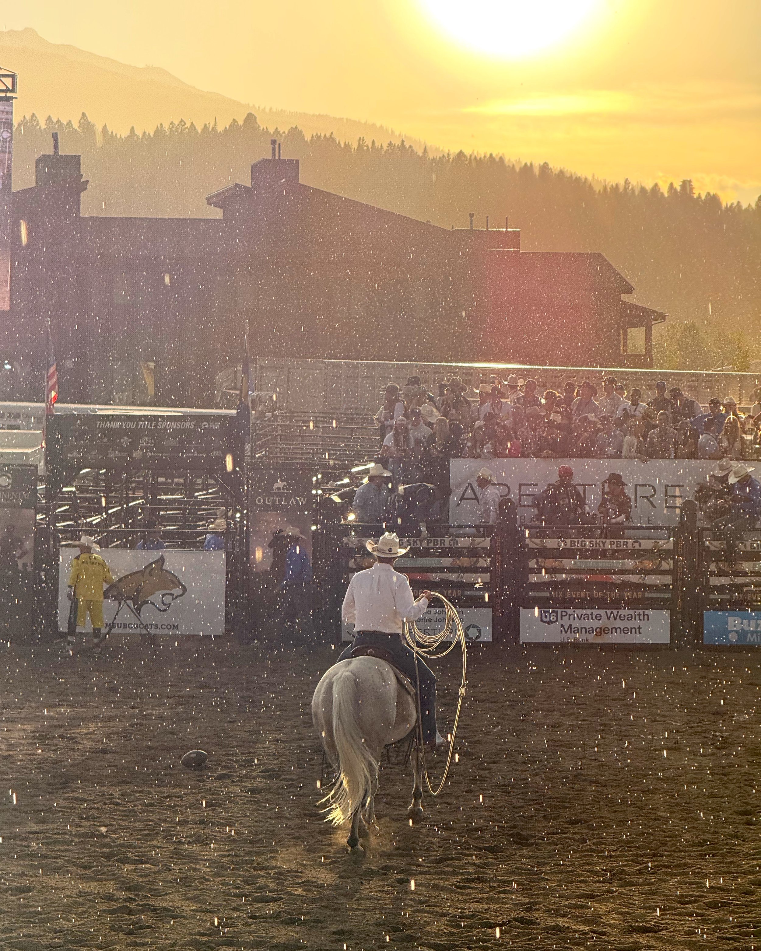 Cowboy riding a white horse on dirt race track at sunset, with spectators in the stands and a backdrop of mountains and trees at the PBR Rodeo in Big Sky, Montana.