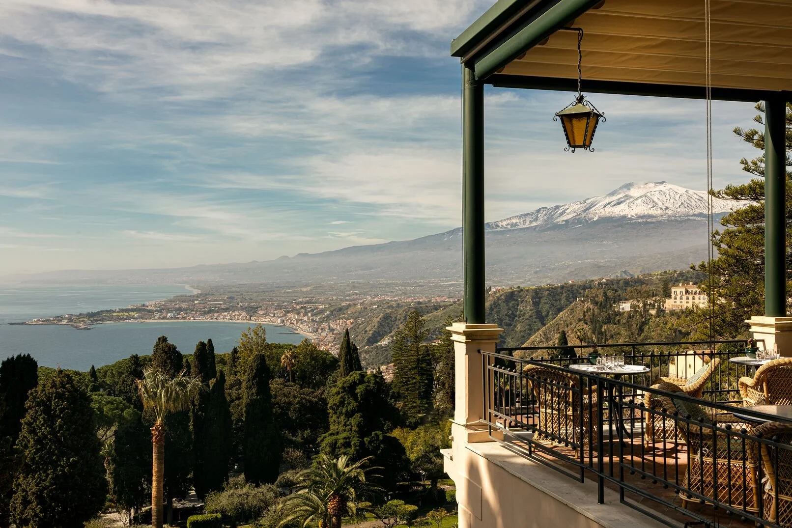A scenic view from a balcony overlooking a coastline, lush trees, and a snow-capped mountain in the distance, with part of a covered patio, a hanging lantern, and outdoor dining furniture in the foreground.
