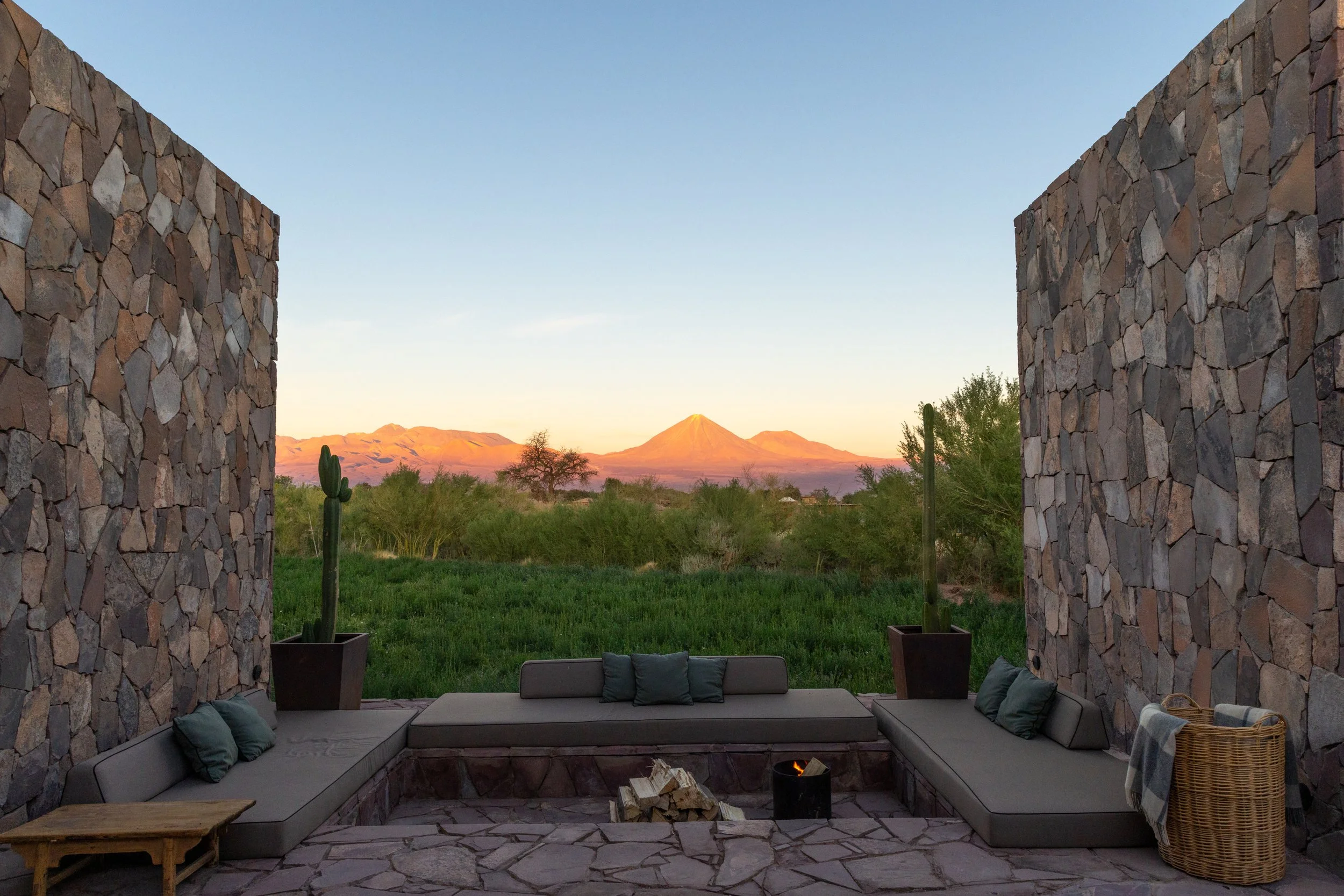 An outdoor seating area with stone walls and cushions, overlooking a desert landscape with cacti and mountains in the distance, during sunset.