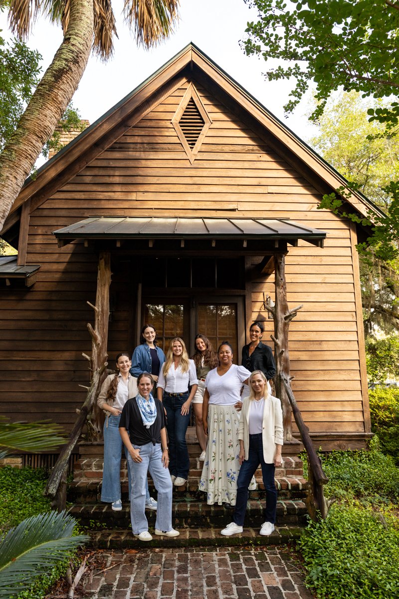 A group of nine women standing on the brick steps in front of a wooden house with trees and greenery around.