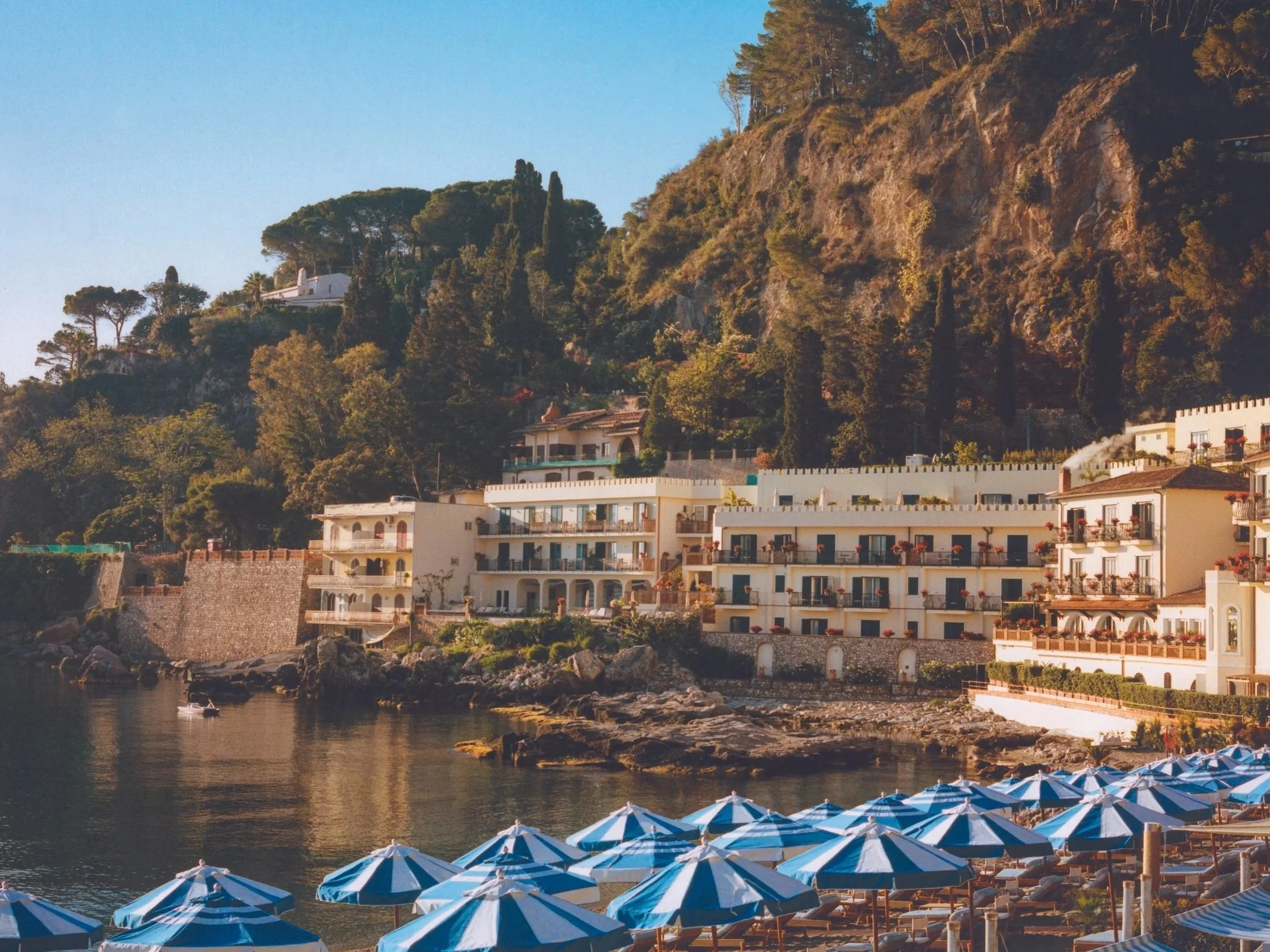 Beach scene, in Taormina, Sicily with blue and white striped umbrellas along the shoreline, with luxury hillside houses and lush green trees on a rocky hill in the background, under a clear blue sky.