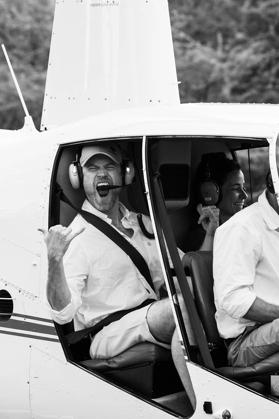Two people sitting in the cockpit of a helicopter, wearing headsets. The man in the front is making a rock and roll hand gesture and shouting. The woman behind him is smiling and holding her headset. The photo is in black and white.