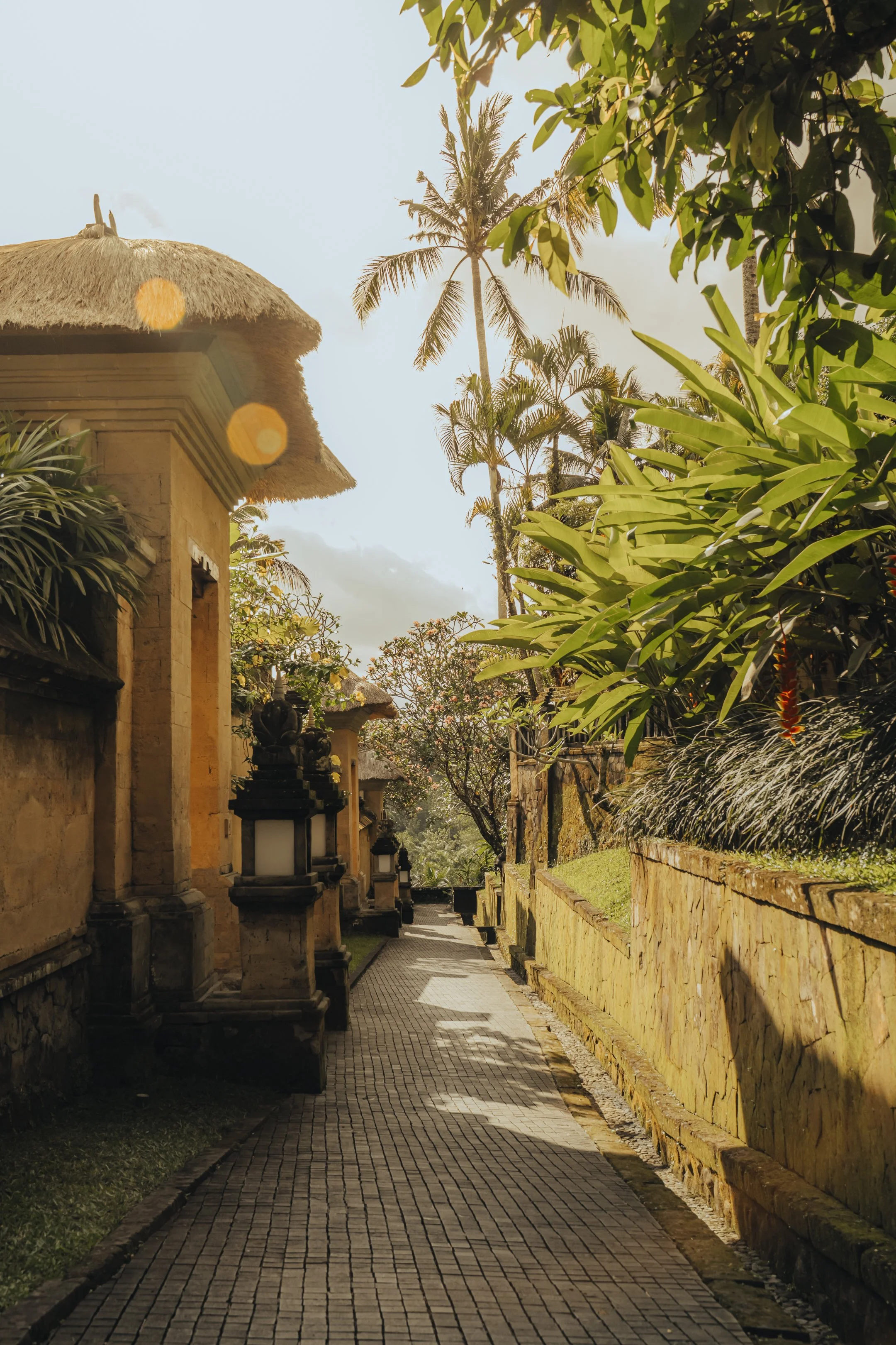 A stone pathway lined with traditional Balinese temple lamps and walls, with lush green trees and palms on both sides under a partly cloudy sky.