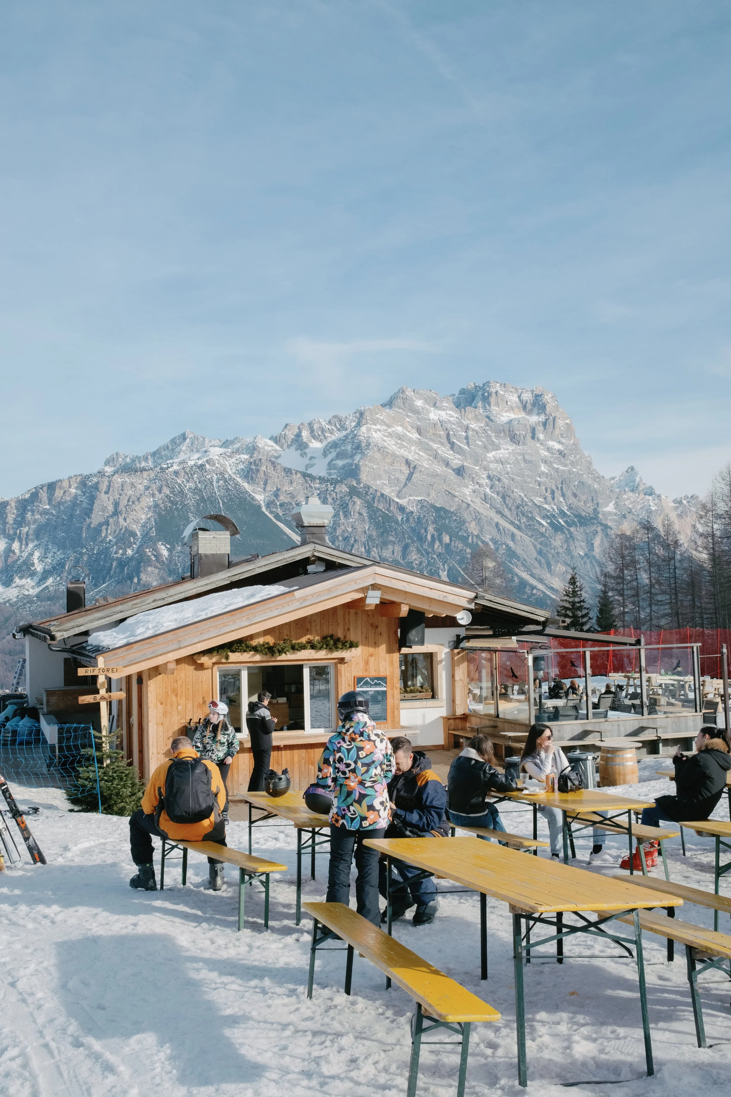 Outdoor seating area at a mountain cafe with people, snow on the ground, wooden tables and benches, a wooden building, and snow-capped mountains in the background.