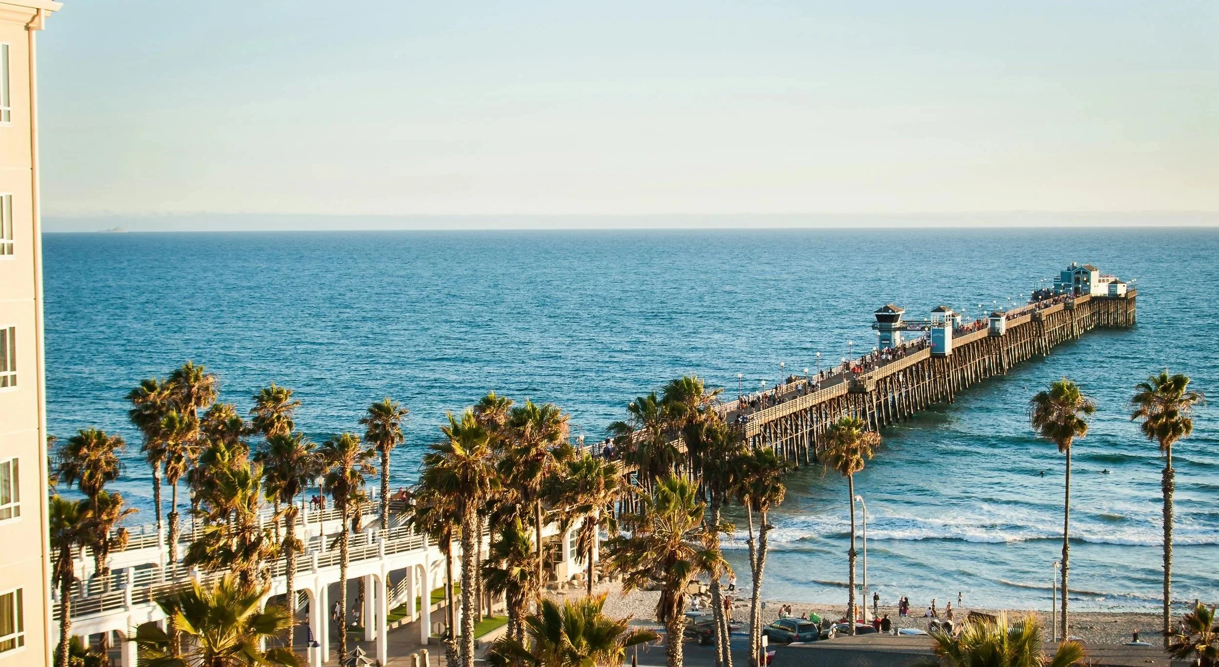 Beachside pier with people walking, lined with palm trees and adjacent buildings, extending into the ocean.