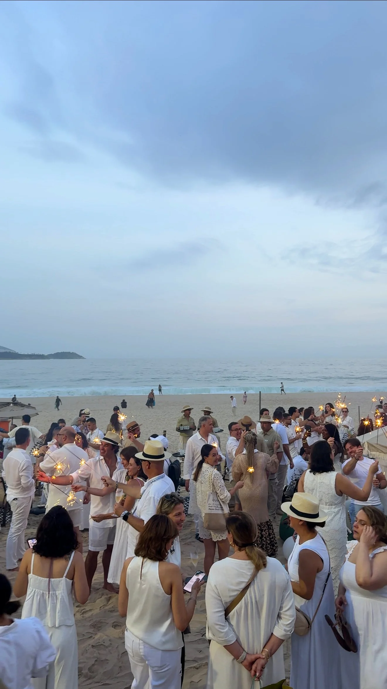 A large group of people dressed in white attending a celebration or gathering on a beach, with some holding sparklers. The ocean and sky are visible in the background.
