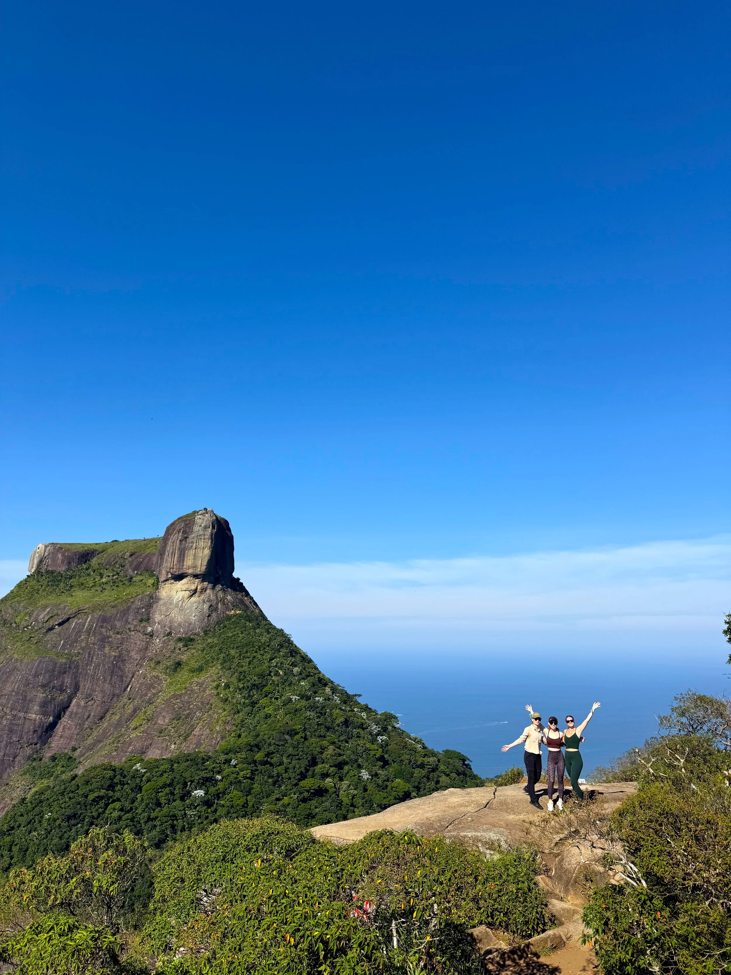 Three people standing on a large rock with a mountain and blue sky in the background, smiling and posing with arms raised. They are at a view point overlooking Rio.