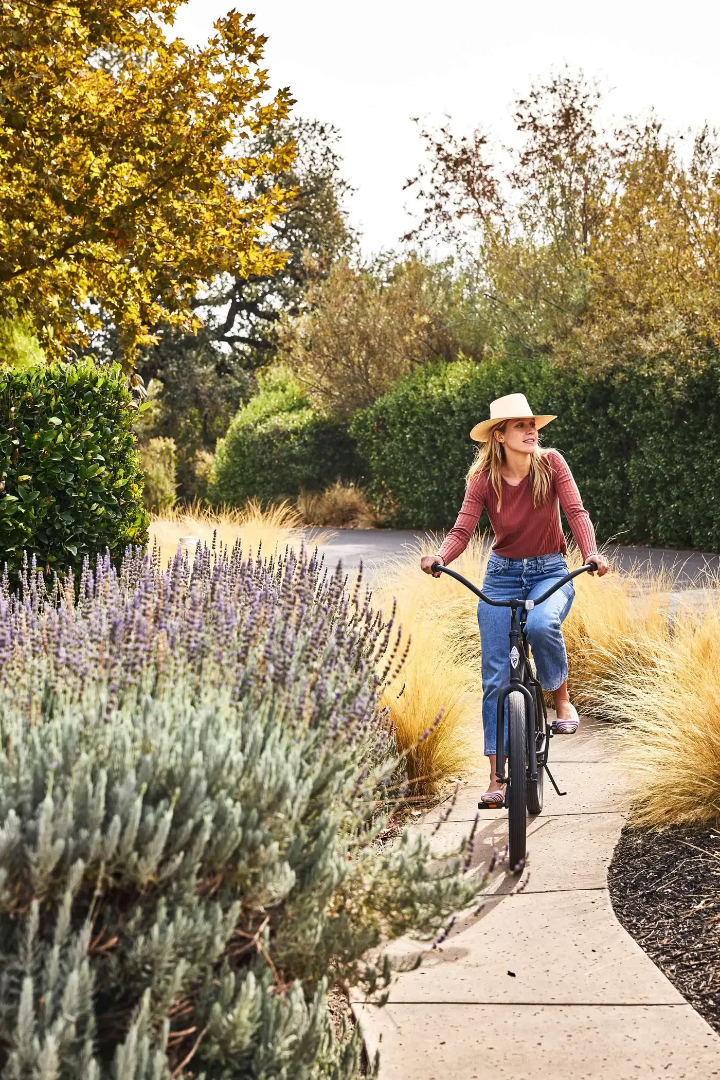 A woman wearing a wide-brimmed hat and weathered jeans is riding a bicycle on a garden path surrounded by lush shrubs and tall ornamental grasses.