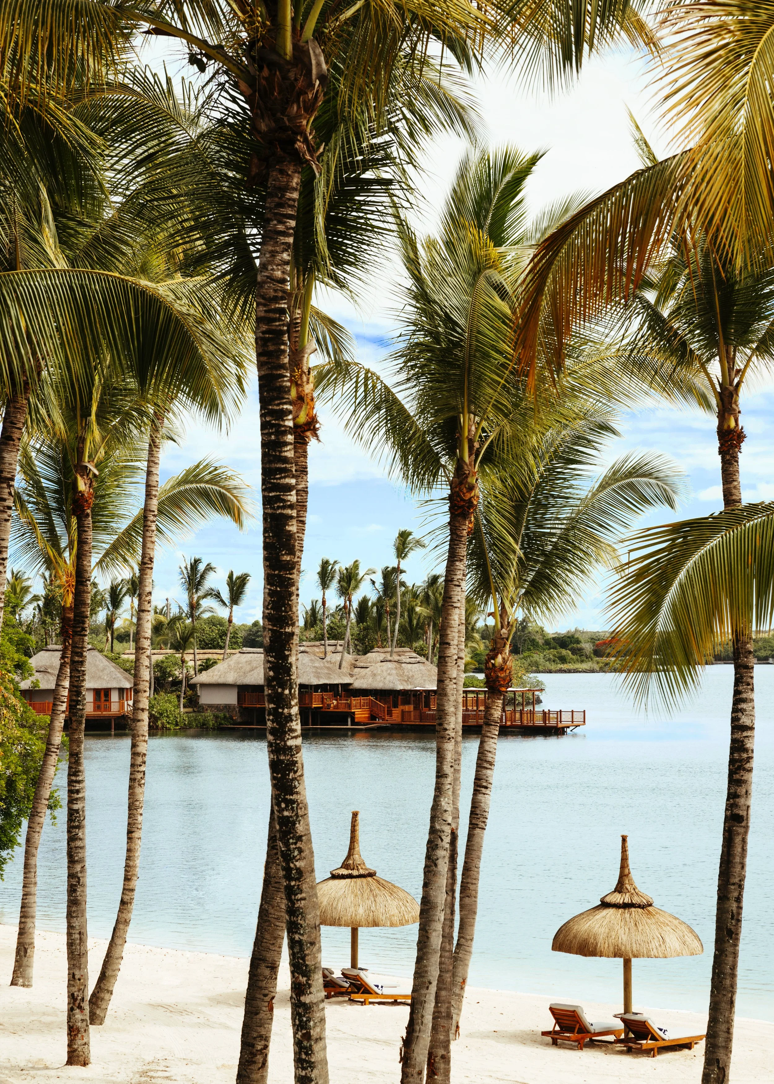 Tropical beach with palm trees, thatched umbrellas, and lounge chairs near the water, with overwater bungalows in the background under a partly cloudy sky.