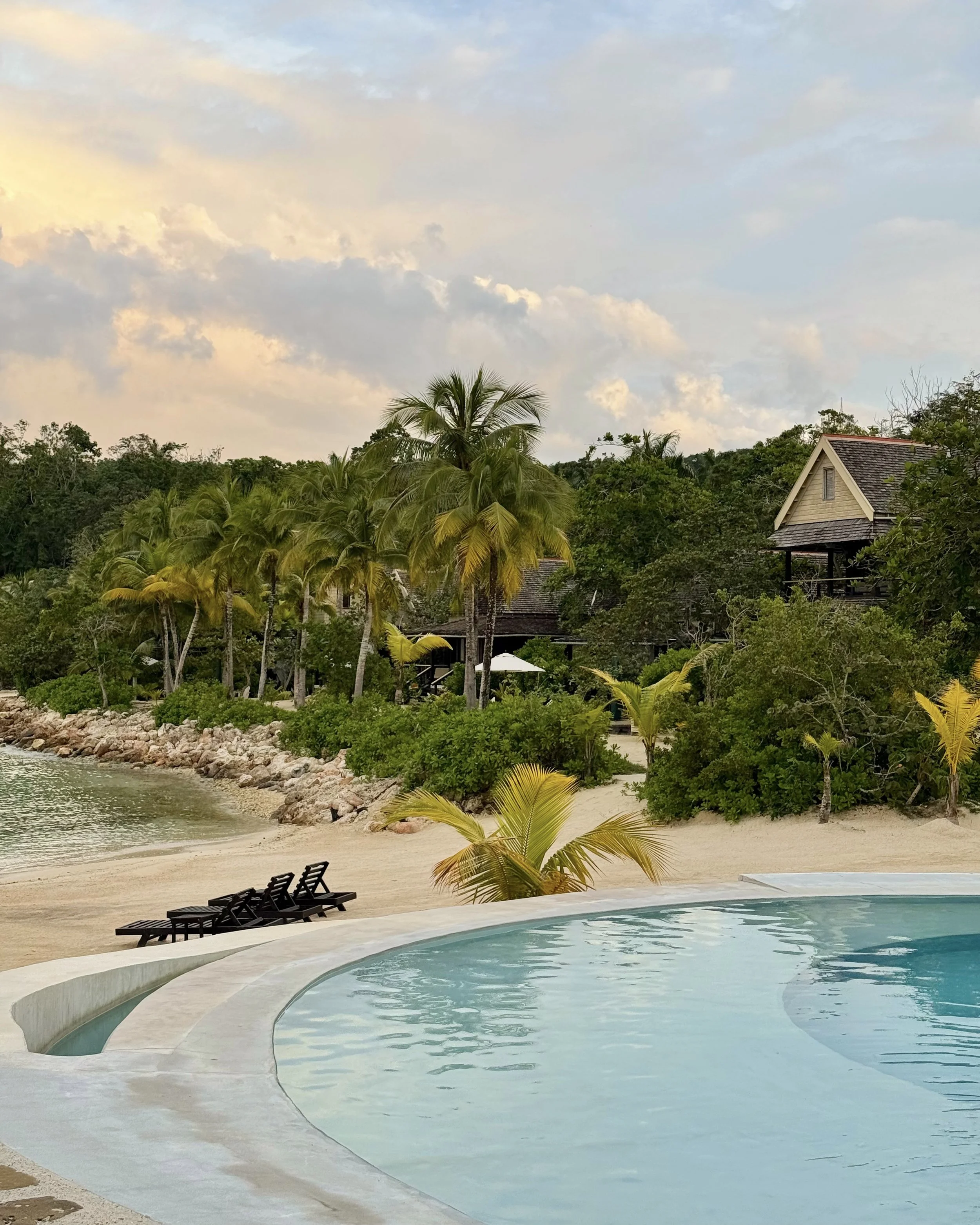 A serene tropical resort scene with a curved swimming pool in the foreground, lounge chairs, a sandy beach with lush green palm trees, and a house among the trees, all under a partly cloudy sky at GoldenEye Resort in Jamaica.