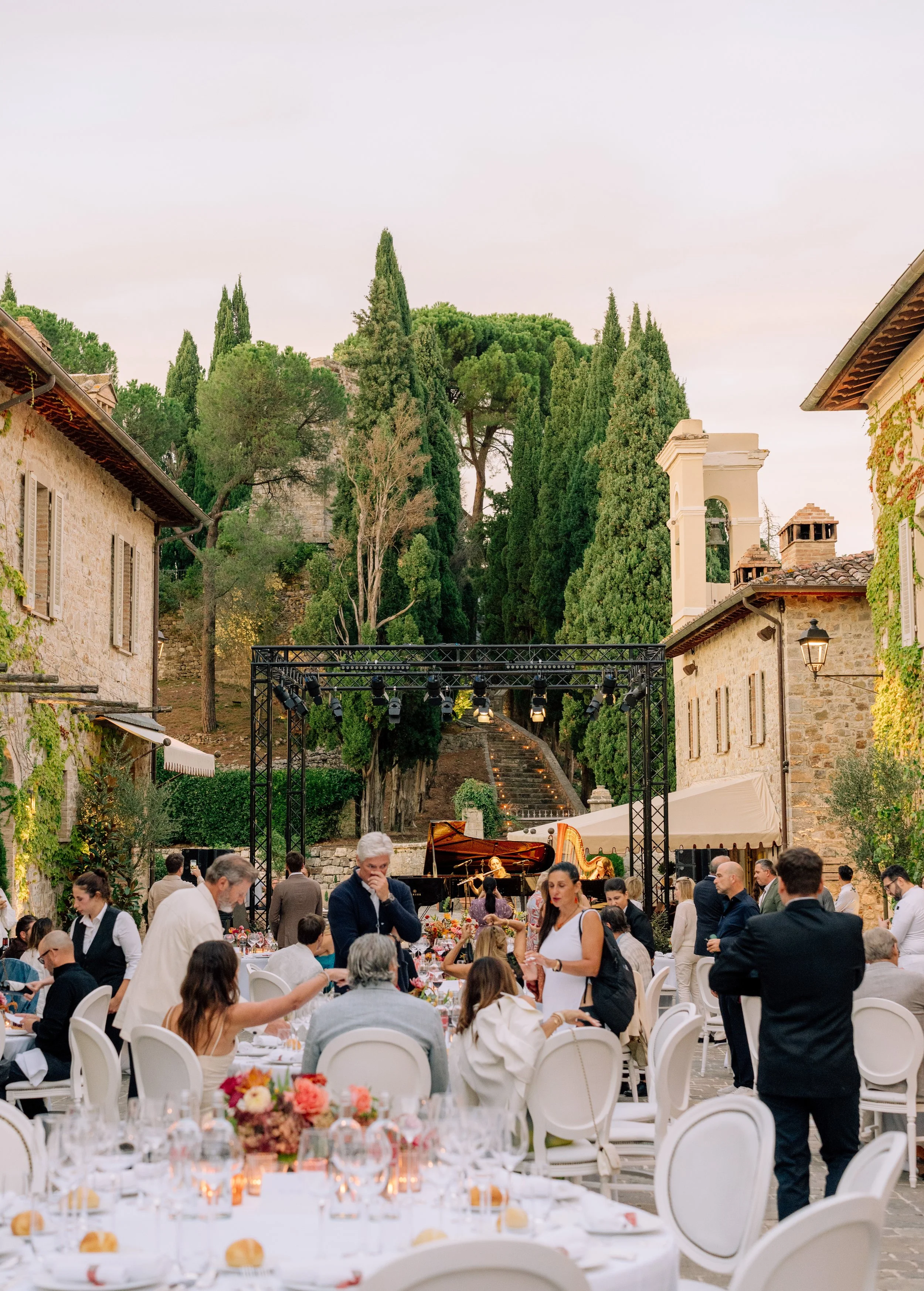 A lively outdoor event with numerous guests seated at tables, enjoying a meal, with a piano performance on stage, set in a charming courtyard surrounded by stone buildings and lush tall trees.