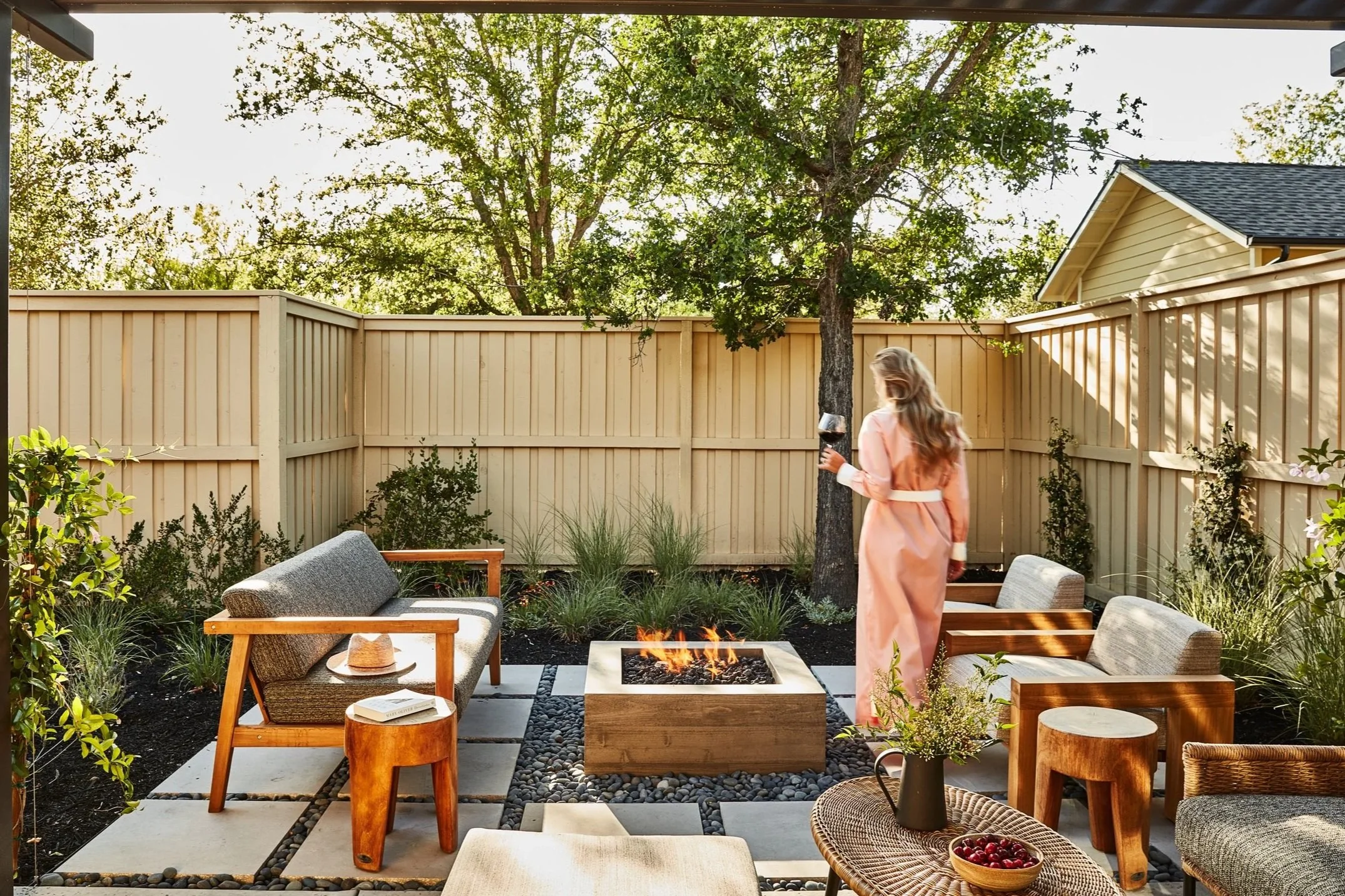 A backyard patio with seating and a fire pit, surrounded by a wooden fence and trees, with a woman in a pink coat holding a wine glass, during daylight.