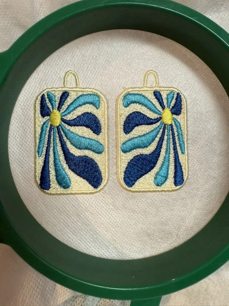 Embroidery of a pair of blue, teal, and navy nail polish bottles with abstract designs, placed inside a green embroidery hoop.
