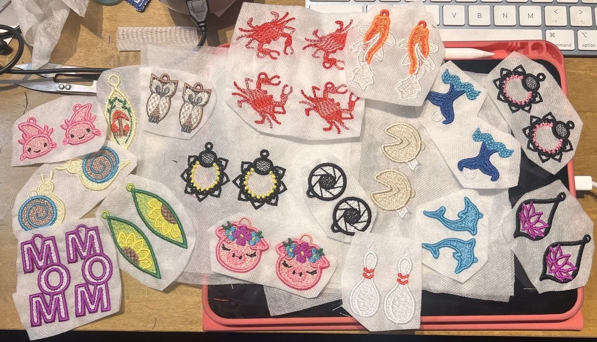 Collection of embroidered earrings with various colorful designs, including animals, moons, flowers, food, and mythical creatures, spread out on a workspace.
